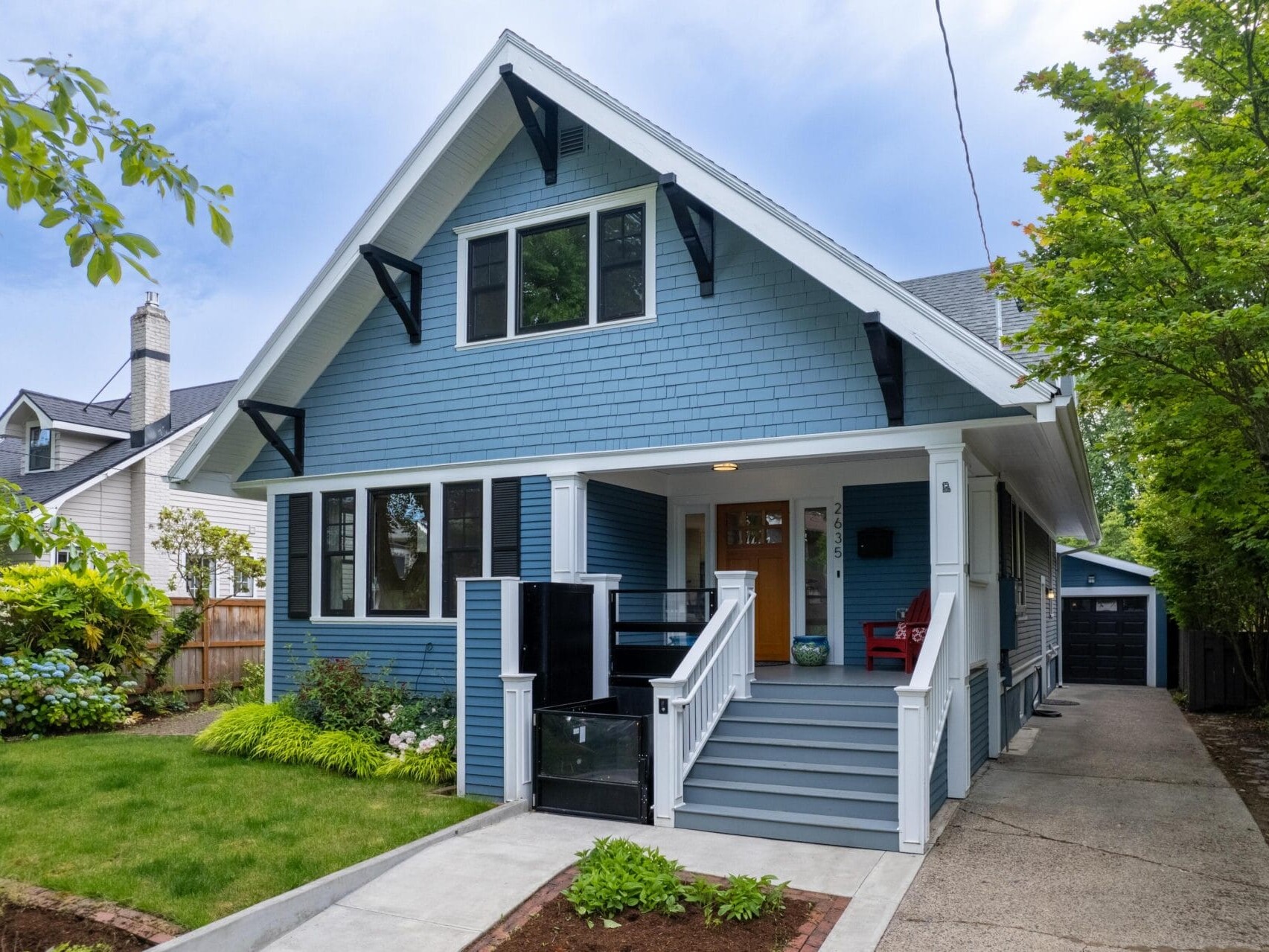A charming blue Craftsman-style house with a gabled roof and white accents. It features a welcoming front porch with red chairs, a well-kept front yard, and a driveway leading to a detached garage surrounded by trees and shrubs.