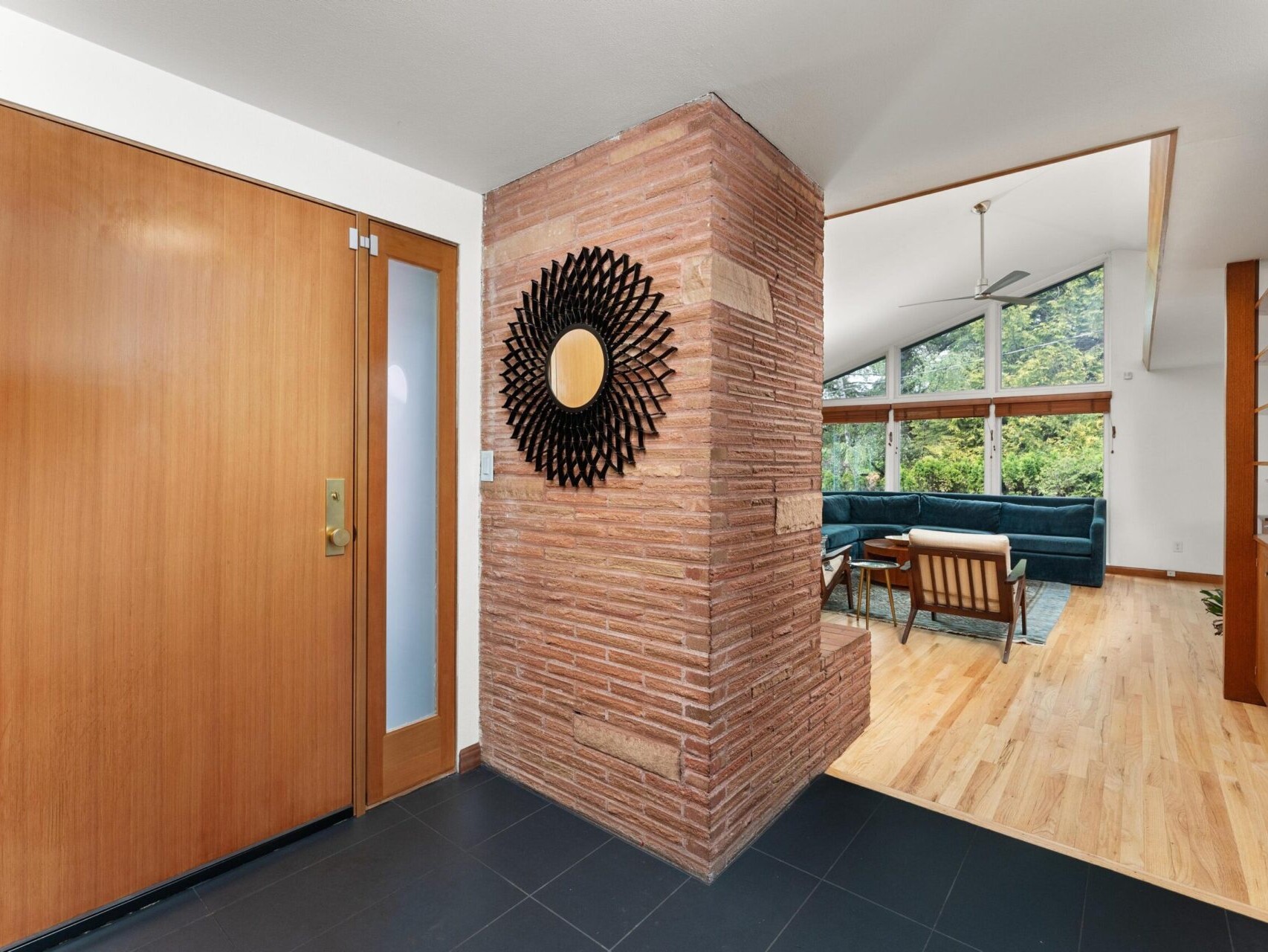 A modern interior features a brick accent wall with a decorative mirror, leading to a living room. The living room has large windows showing trees outside, a blue sofa, wooden floors, and an open ceiling with a fan.