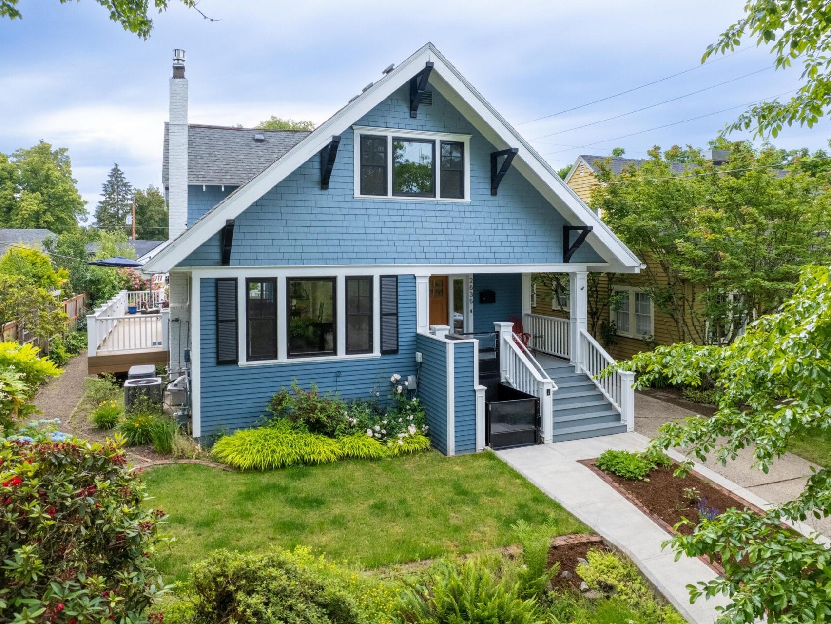 A charming blue two-story house with white trim, featuring a front porch and a well-maintained garden. The lawn is bordered by various shrubs and trees. A paved path leads to the steps of the house, surrounded by greenery and neighboring homes.