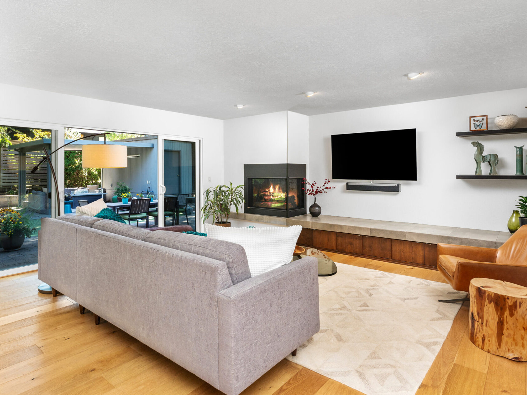 A modern living room with a grey sofa, a leather chair, and a wooden table. A fireplace and a large TV are on the wall. Sliding glass doors lead to an outdoor patio with plants. Shelves hold decorative items, and the floor is light wood.