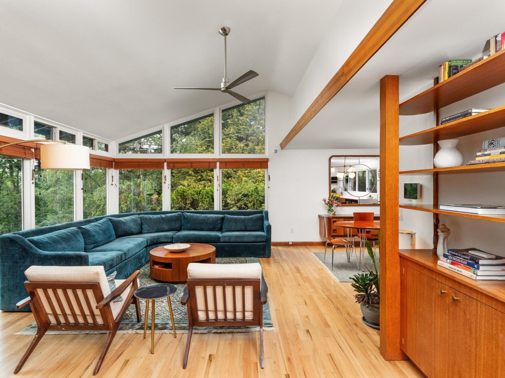 A mid-century modern living room featuring a large, blue sectional sofa and two wooden chairs with white cushions. The room has large windows, a wooden bookshelf, and a ceiling fan, with views of greenery outside.