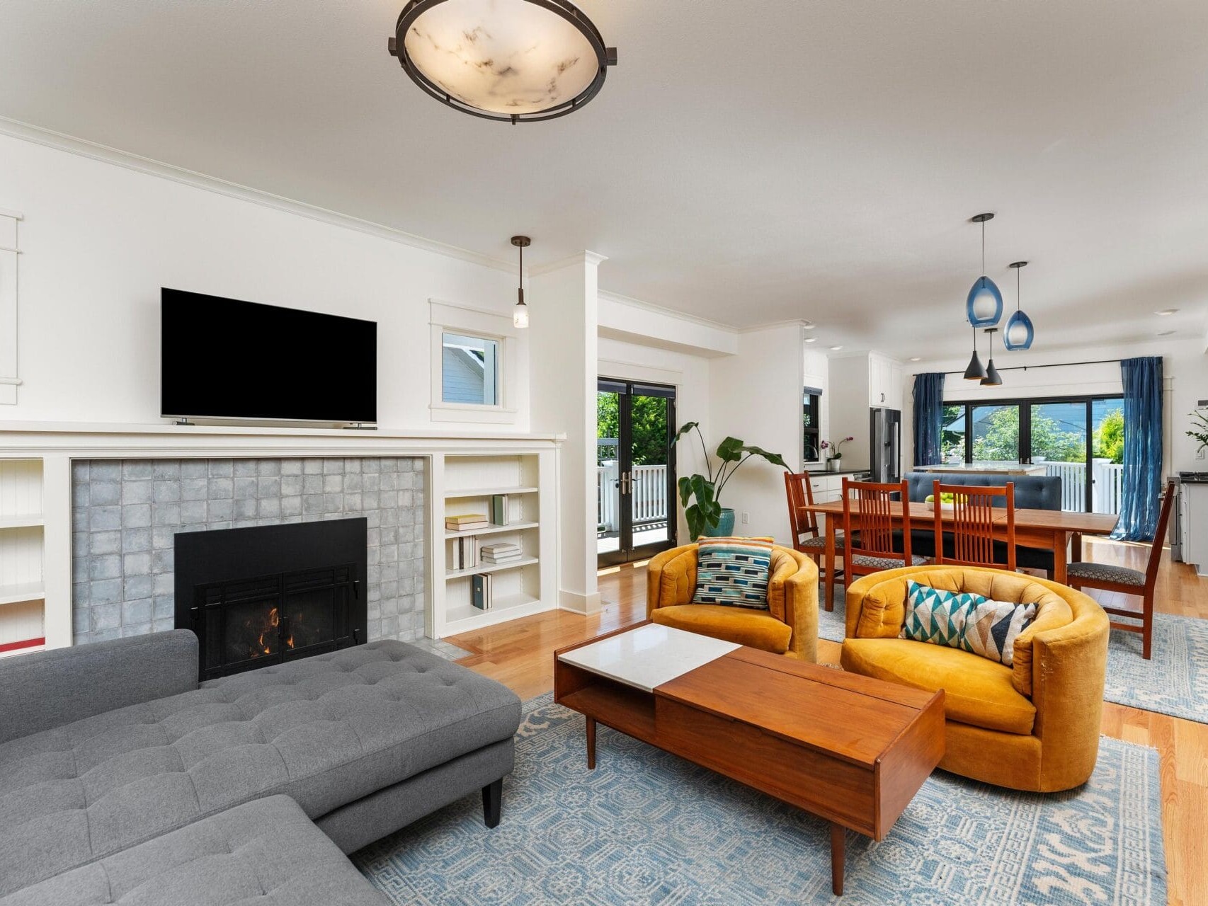 A modern living room with a gray sectional sofa, two yellow armchairs, and a wooden coffee table. A fireplace with a TV above it is flanked by bookshelves. A dining area and large windows with blue curtains are visible in the background.