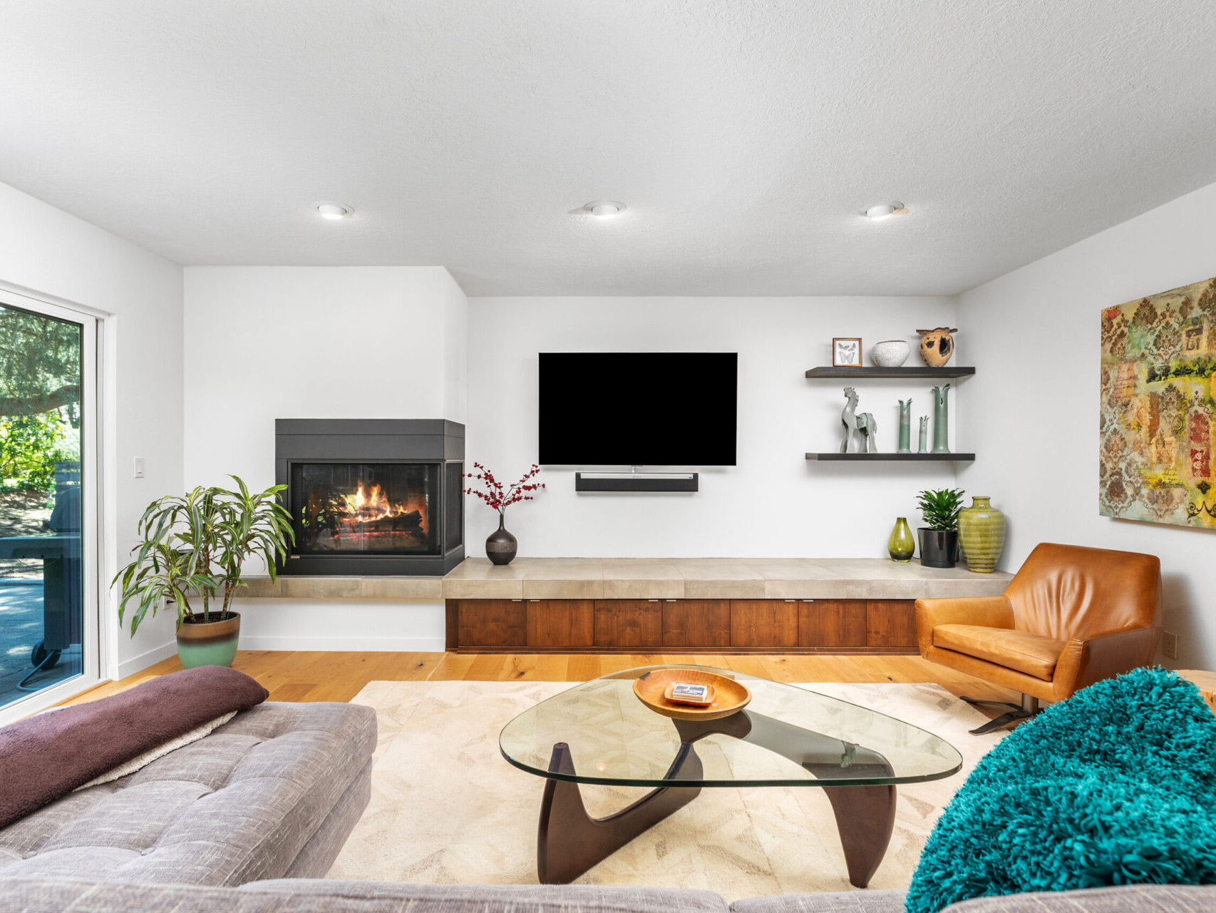 A modern living room with a wall-mounted TV above a fireplace. The room features a brown leather chair, gray couch, glass coffee table, and decorative items on shelves. Large windows brighten the space and a colorful painting hangs on the wall.