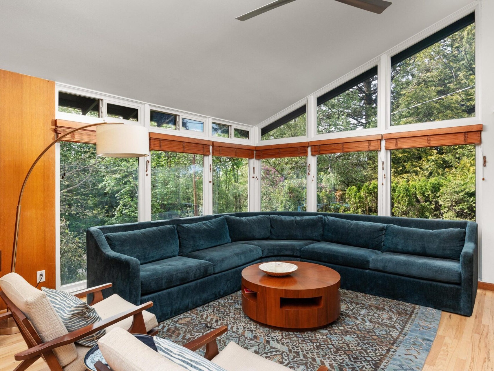 A cozy living room with a large blue sectional sofa arranged in a semi-circle around a round wooden coffee table. Floor-to-ceiling windows showcase a lush green view outside. The room features wooden accents and a patterned rug.