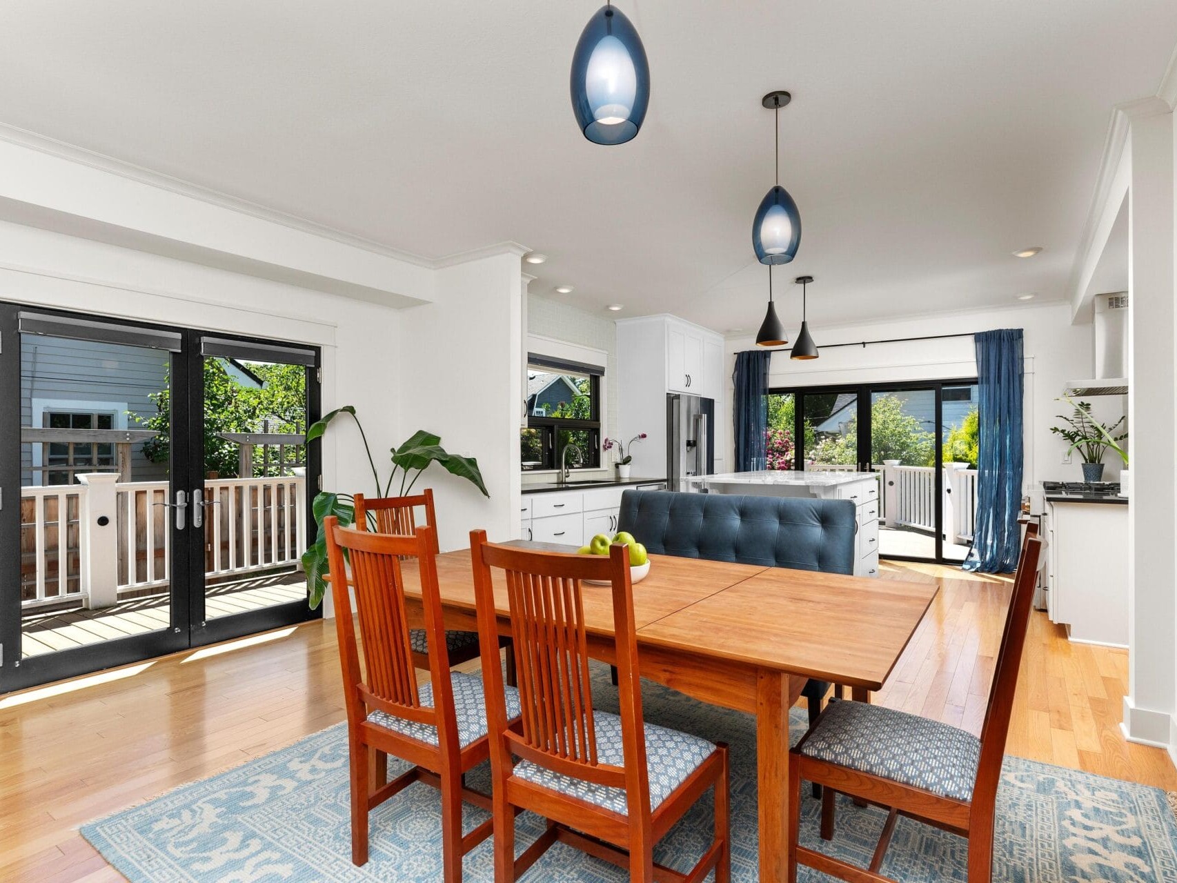 A bright dining area with a wooden table and chairs on a blue rug. The room has large glass doors leading to a deck with greenery outside. The kitchen in the background features white cabinets and modern pendant lights.