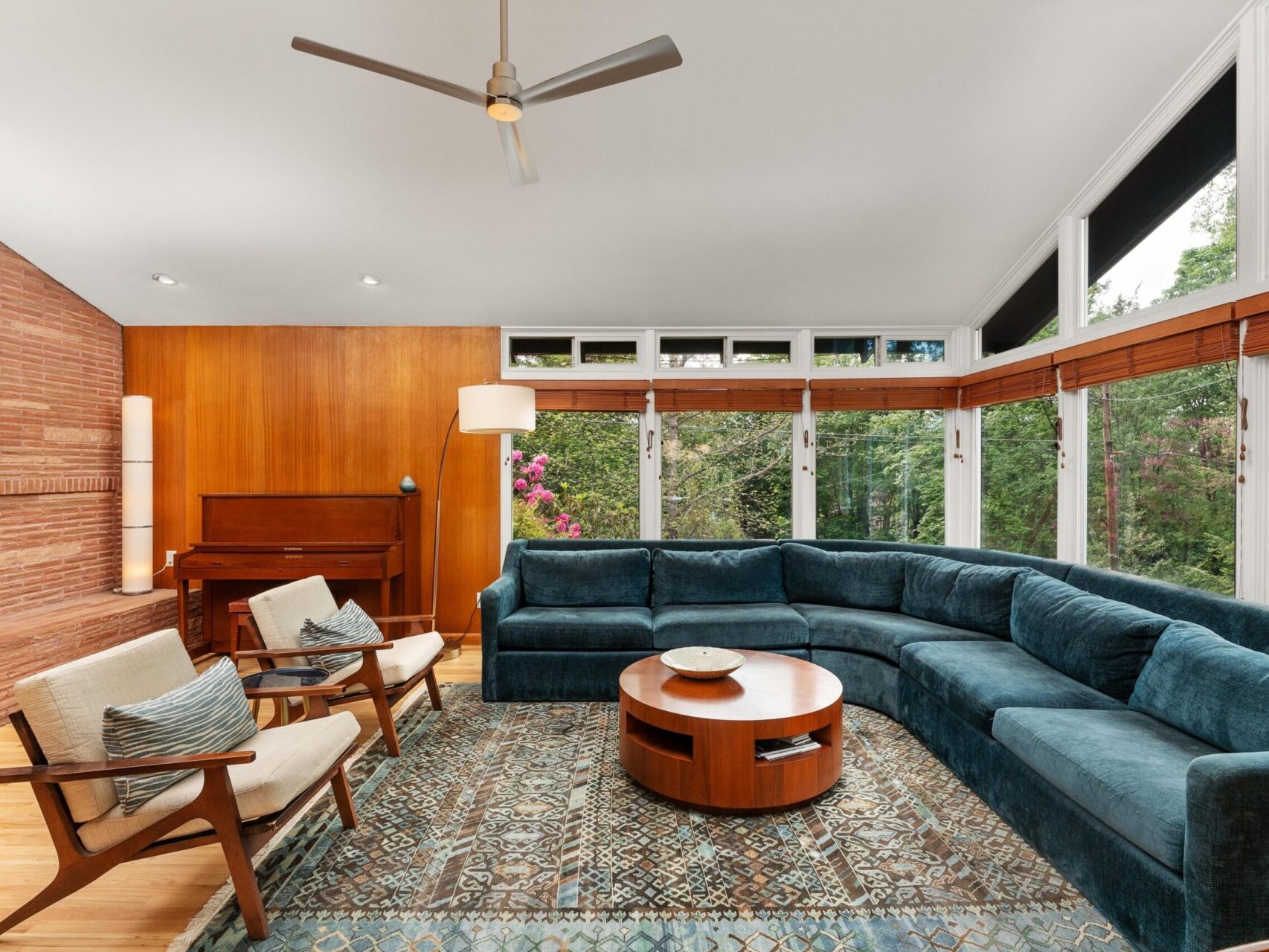A modern living room featuring a large curved blue sofa, two cushioned chairs, and a wooden coffee table on a patterned rug. A piano stands against the wood-paneled wall, and large windows offer views of greenery outside.