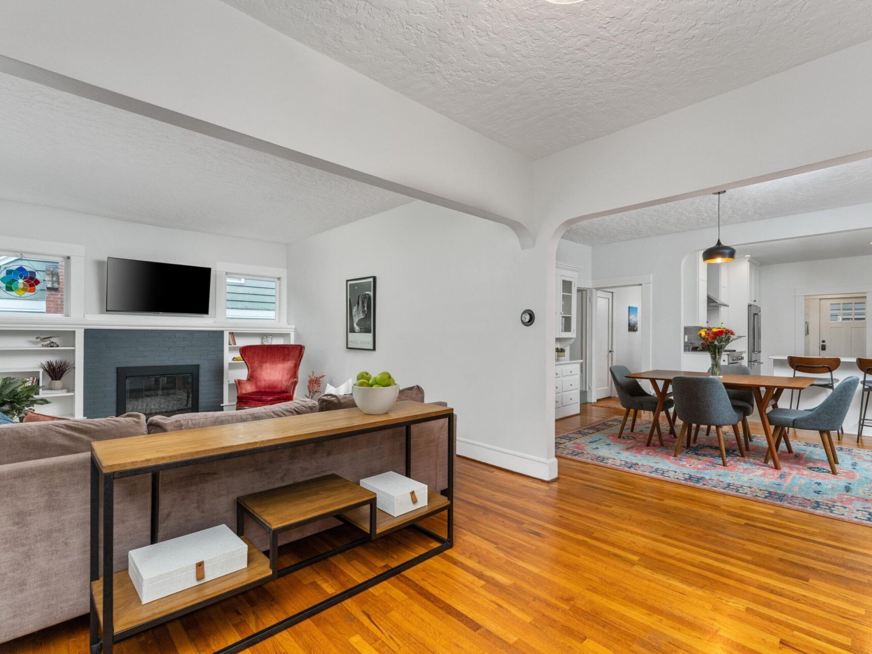 Open living room with wooden floors, featuring a gray sofa and a red chair facing a fireplace with a TV. A dining area with a table and chairs is visible, along with a kitchen in the background. Shelves and a side table display decorative items.