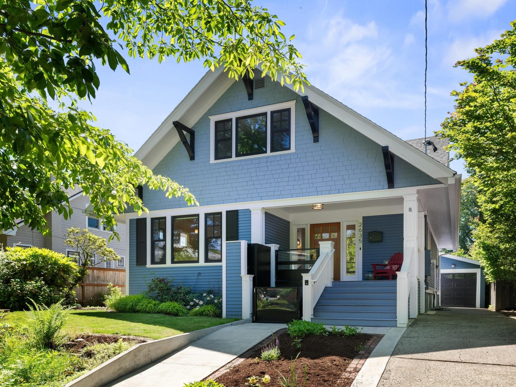 A charming blue house with a pointed roof and white trim, surrounded by lush greenery. It features a front porch with red chairs, a well-maintained lawn, and a driveway leading to a detached garage.