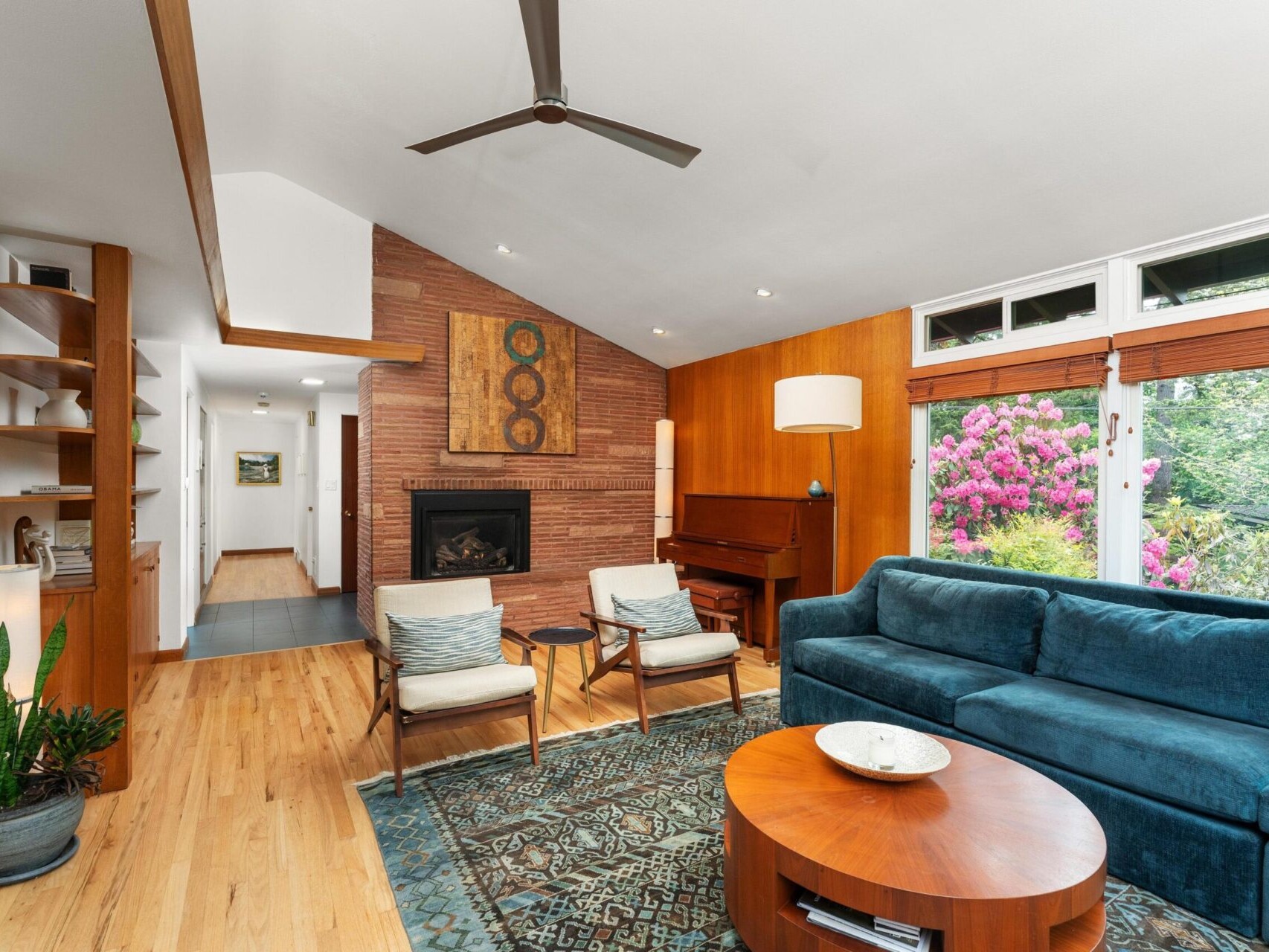 A cozy living room with a blue sofa, two chairs, a round coffee table, and a brick fireplace. A grand piano sits nearby. Large windows overlook a garden with blooming flowers. Ceiling fan and wooden floor add warmth to the space.