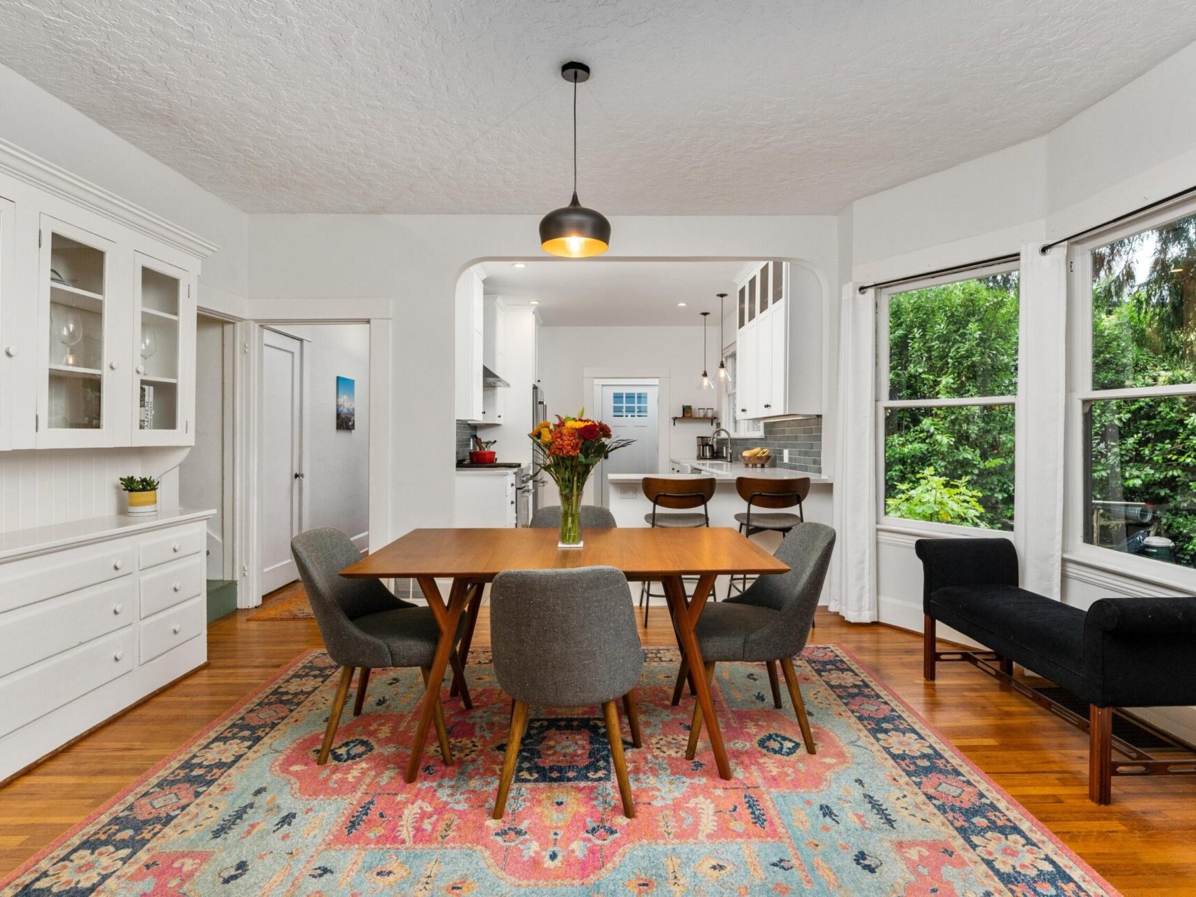 A dining room with a wooden table and six chairs on a colorful patterned rug. A vase with flowers is on the table. A pendant light hangs above. The room opens to a kitchen with stools at a counter. Large windows show green trees outside.