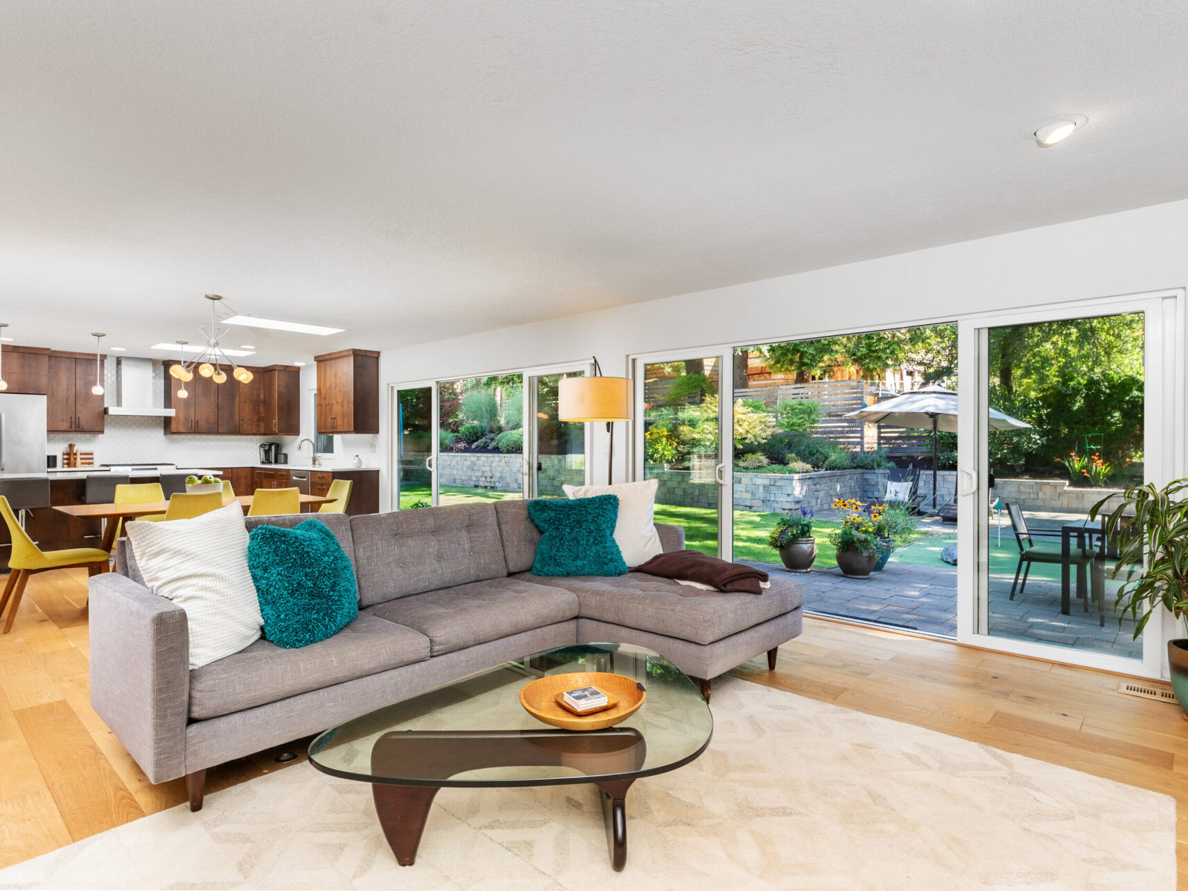 A modern living room with a gray L-shaped sofa, teal and white pillows, and a glass coffee table. Large windows showcase a sunny garden. The open-plan space includes a kitchen with wooden cabinets and a dining table with yellow chairs.