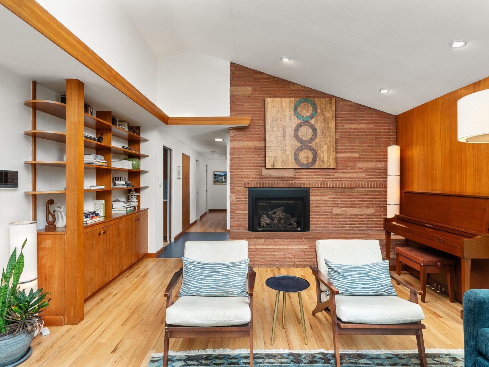 A cozy living room with a wooden piano, a brick fireplace with abstract art above it, wooden shelves filled with books, two cushioned chairs, a small round table, and a potted plant. The design features warm wood tones and a high ceiling.