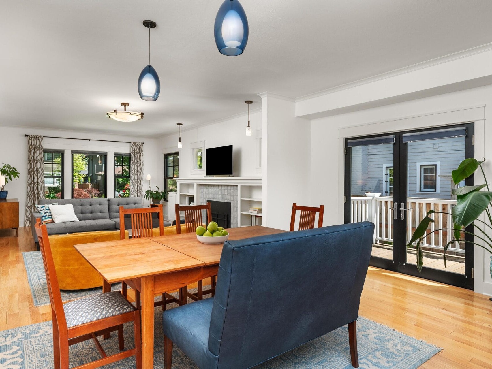 A modern open-plan living and dining area with wooden floors. It features a wooden dining table with chairs, a blue bench, two sofas, a wall-mounted TV above a fireplace, and large glass doors leading to an outdoor area. Hanging lights add elegance.
