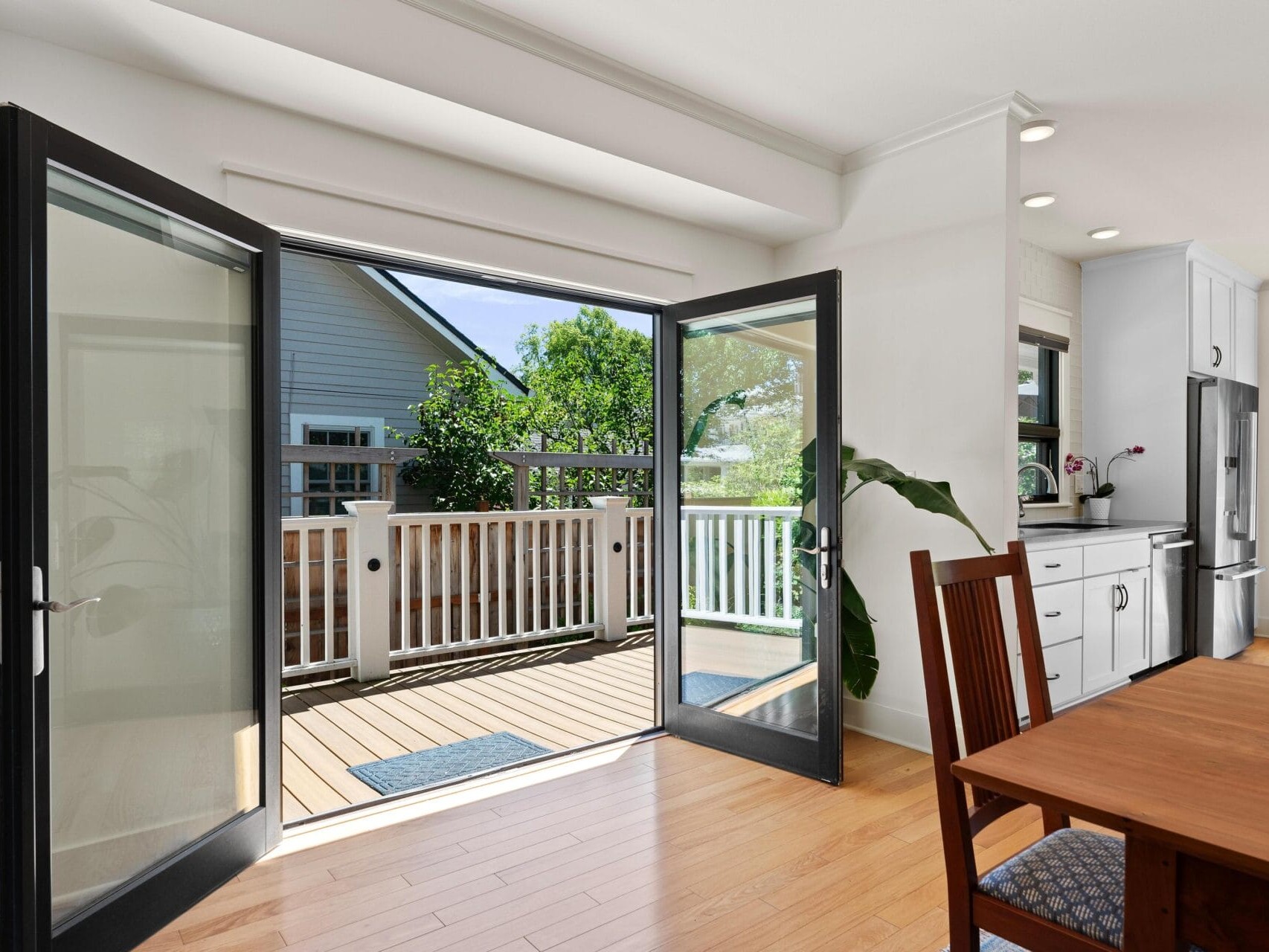 A bright kitchen dining area with wooden floors. French doors open to a wooden deck with a view of a neighboring house and green trees. A wooden dining table and chairs are in the foreground, and the kitchen features white cabinets and stainless steel appliances.