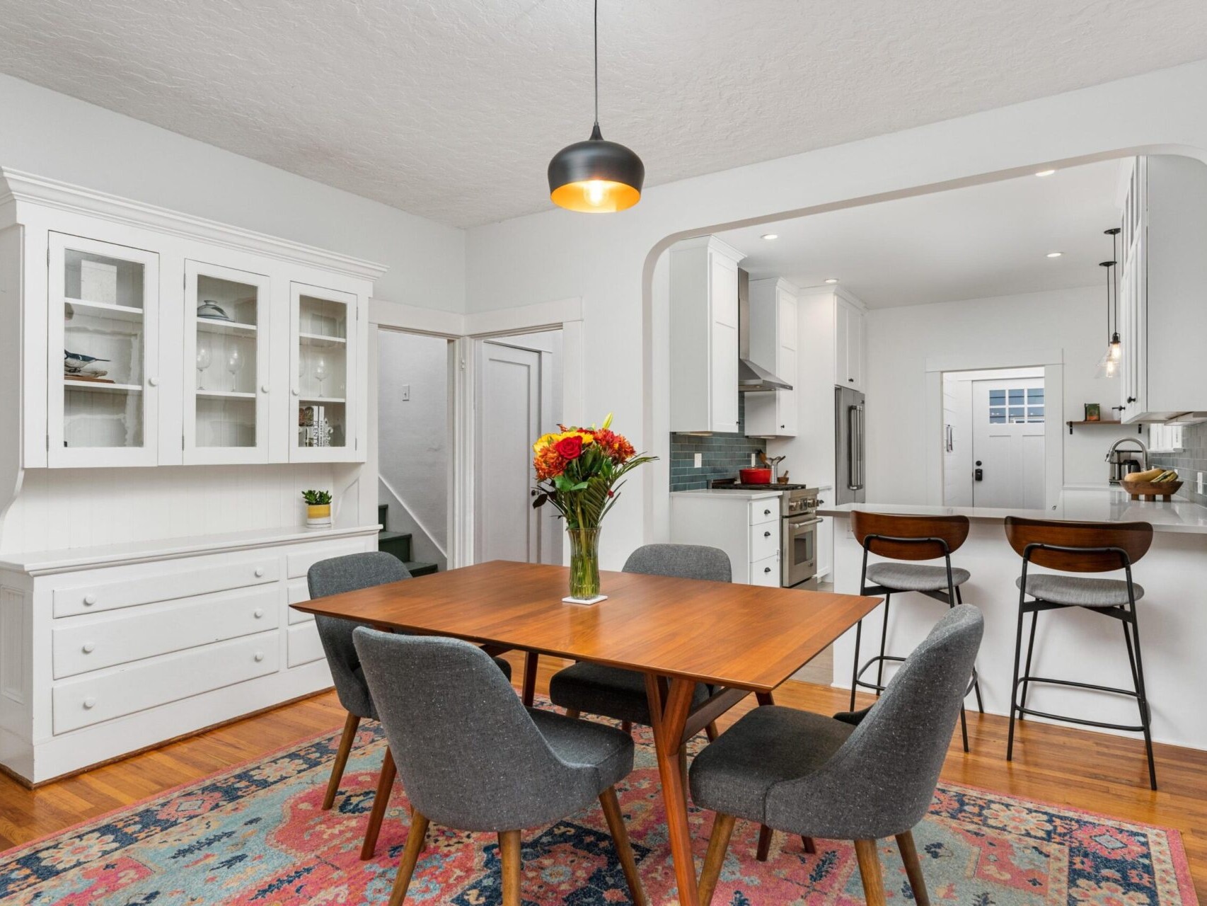 A dining room features a wooden table with six gray upholstered chairs. A vase with flowers is on the table. A colorful rug is on the floor. The background shows a kitchen with white cabinets and a bar area with two stools. A pendant light hangs above.