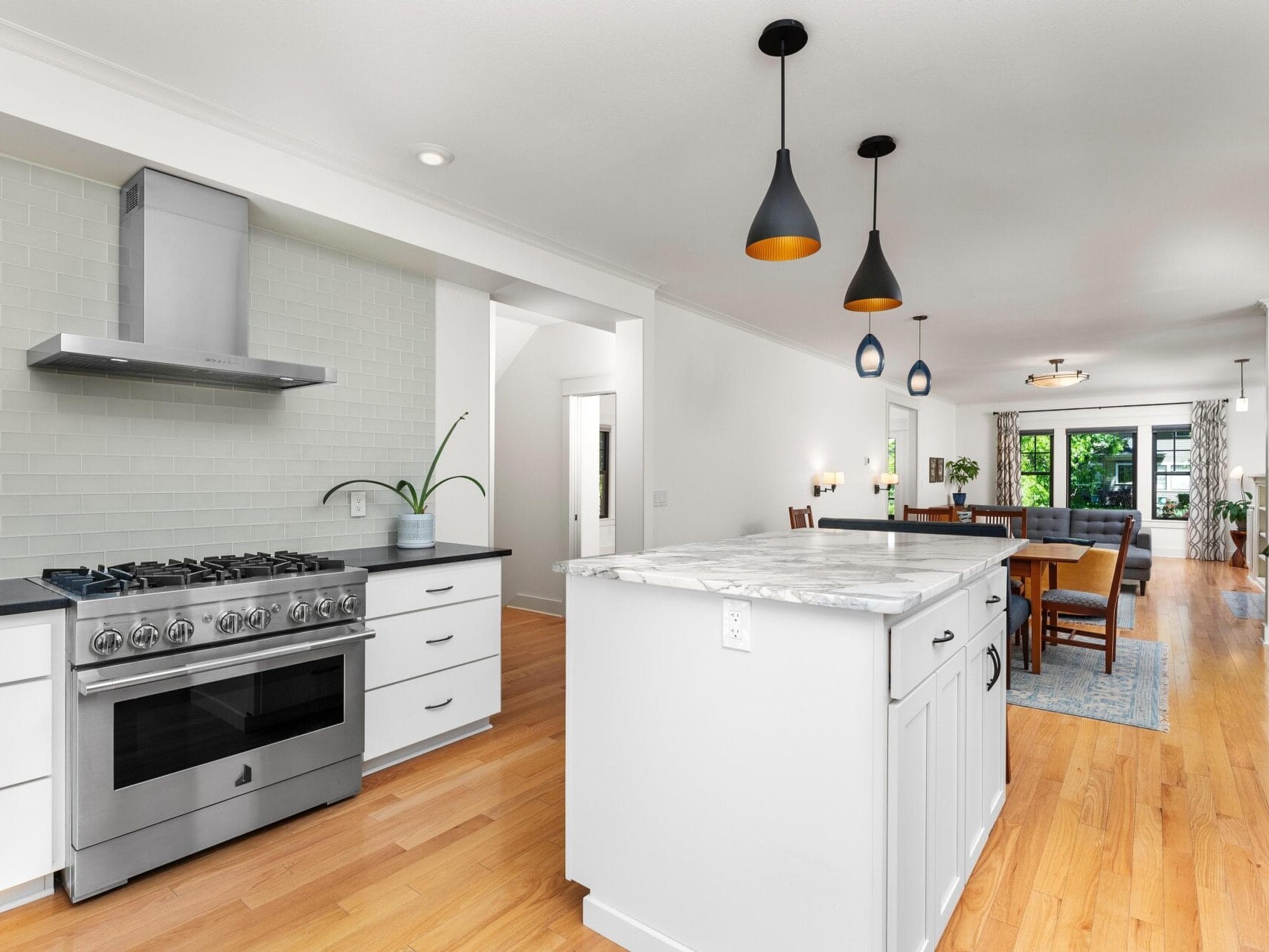 Modern kitchen with a spacious open layout, featuring a stainless steel stove, white cabinets, and a marble island. Pendant lights hang above, and hardwood floors extend to a dining area with a table and chairs near large windows.