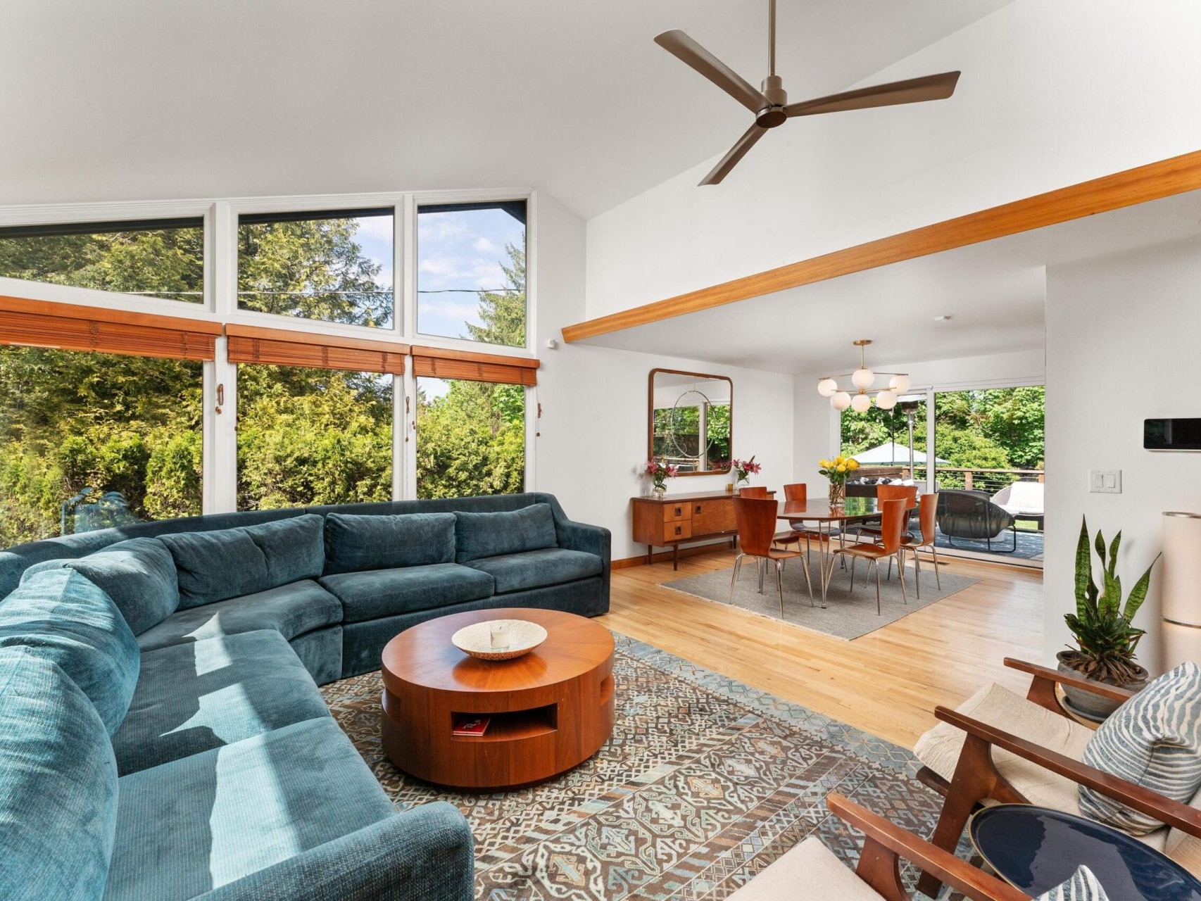 A modern living and dining area with large windows showcasing lush greenery. The room features a blue sectional sofa, a circular wooden coffee table, a ceiling fan, and a dining table with chairs. Decor includes plants and a patterned rug.
