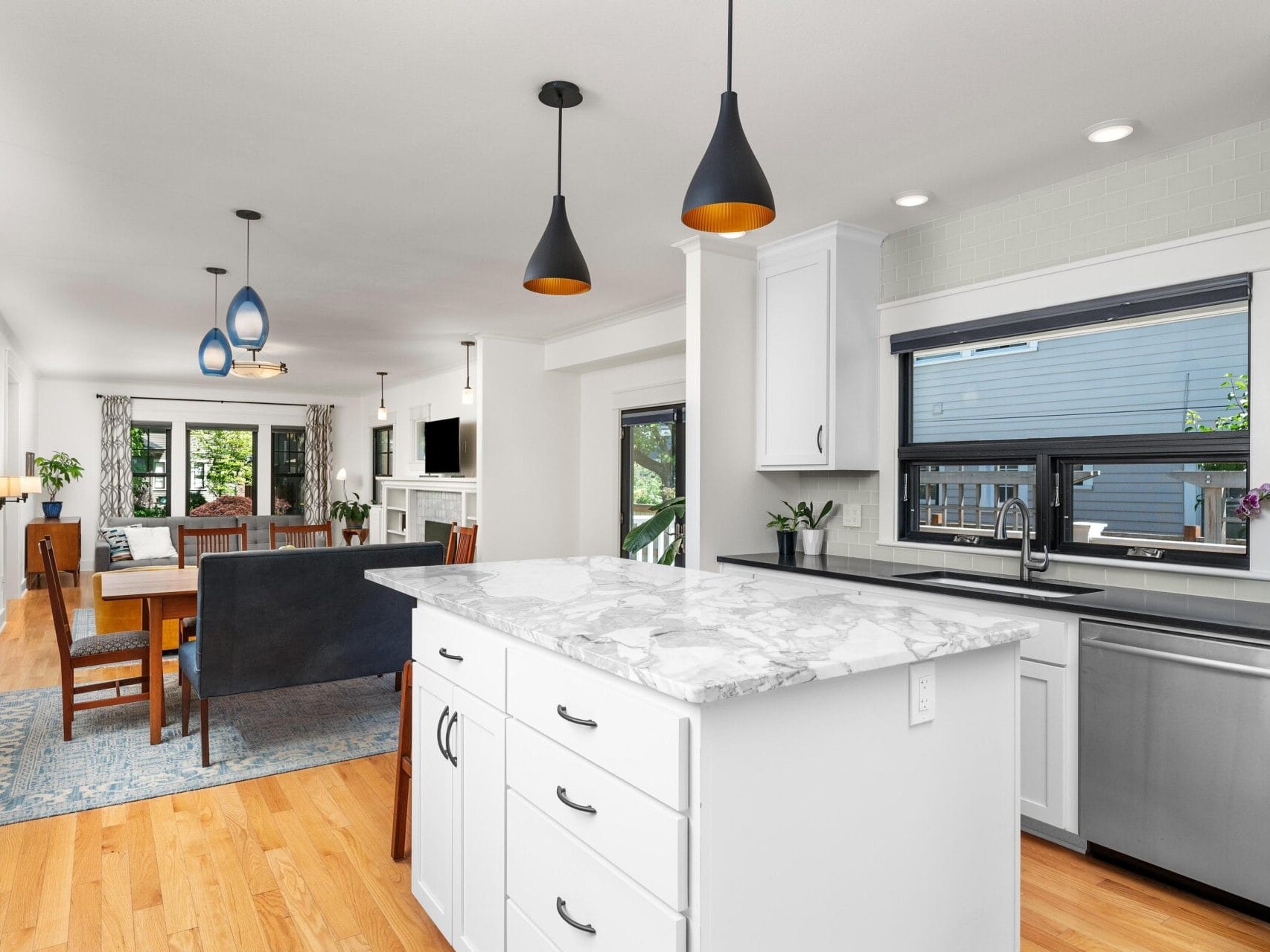 A modern open-plan kitchen and dining area with white cabinets and marble countertops. Three black pendant lights hang above the kitchen island. The dining area features a wooden table and chairs, with a blue rug underneath. Large windows provide natural light.