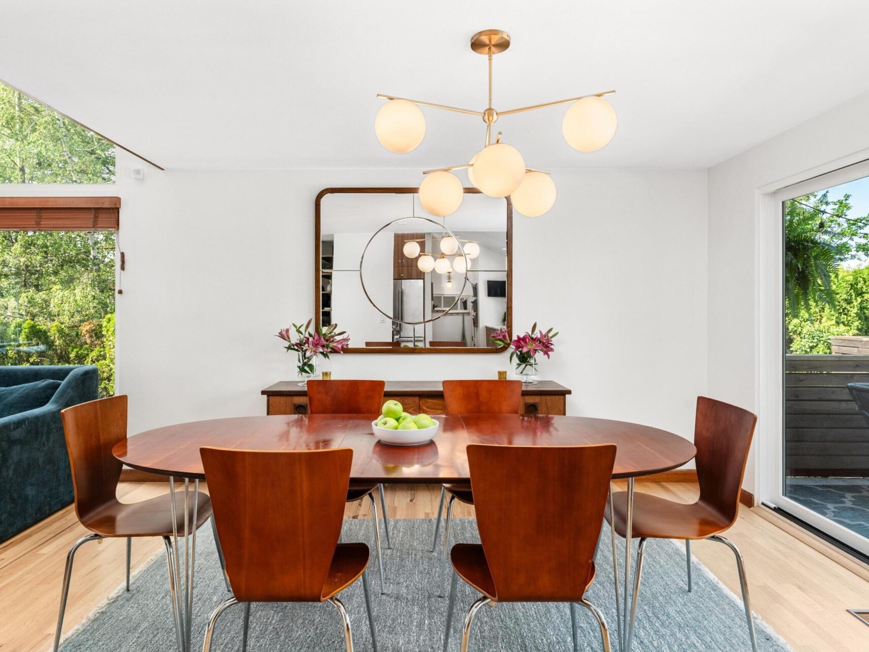 A modern dining room with a wooden oval table and six matching chairs. A sleek chandelier hangs above. A large mirror and vases with flowers adorn a sideboard. Large windows offer a view of greenery, enhancing the bright and airy ambiance.