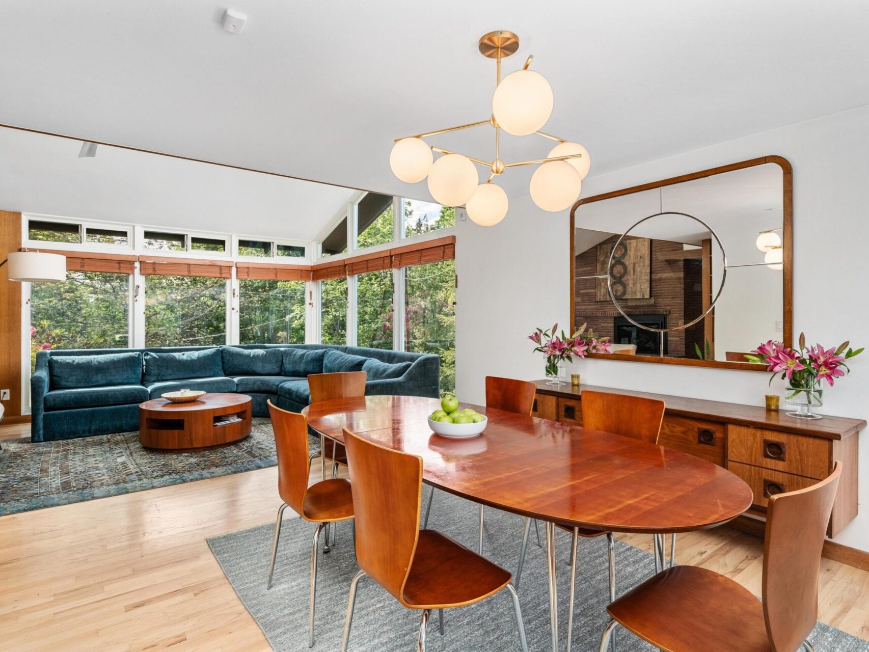 A modern dining room with a wooden oval table and six chairs. A large, contemporary chandelier hangs above. A living area with a teal sectional sofa is visible in the background. The room features large windows and a mirror on one wall.