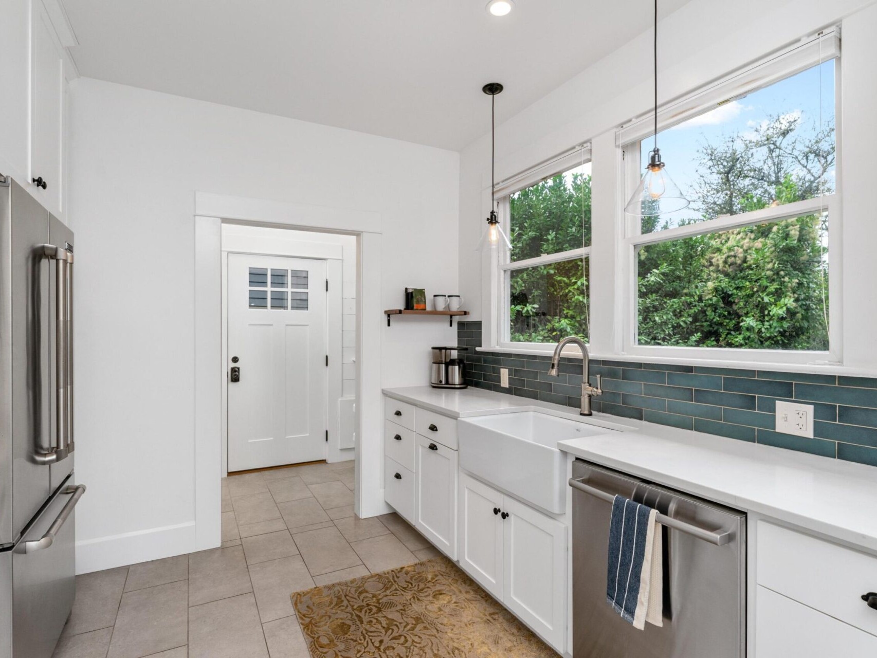 Modern kitchen with white cabinets, stainless steel appliances, and a farmhouse sink. Blue subway tiles line the backsplash. A window above the sink offers a view of greenery outside. Two pendant lights hang from the ceiling, and theres a door nearby.