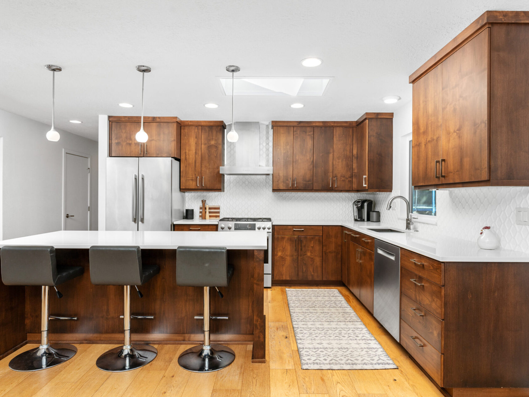 Modern kitchen with wooden cabinetry, a white countertop island with three black barstools, stainless steel appliances, and a patterned rug. Pendant lights hang above the island, and light wood flooring completes the look.