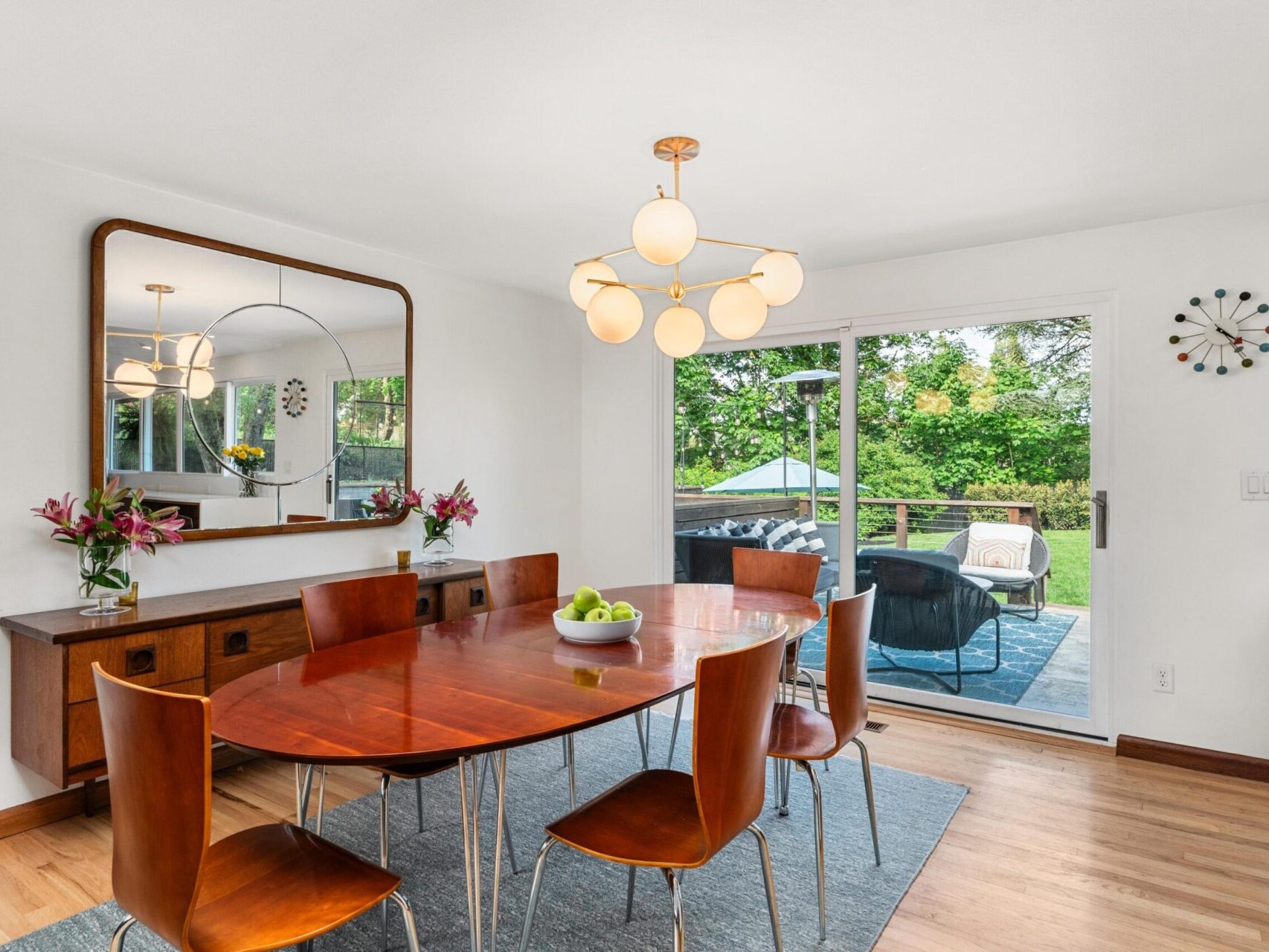 A modern dining room with a wooden table and six chairs. A large mirror hangs above a sideboard with vases of flowers. A chandelier with round bulbs is centered above the table. Sliding glass doors open to a patio with greenery outside.
