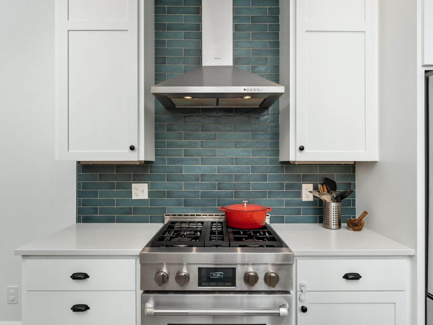 Modern kitchen with a stainless steel stove and range hood, set against a blue subway tile backsplash. A red pot sits on the stove, and nearby are kitchen utensils in a holder. White cabinets with black knobs frame the scene.
