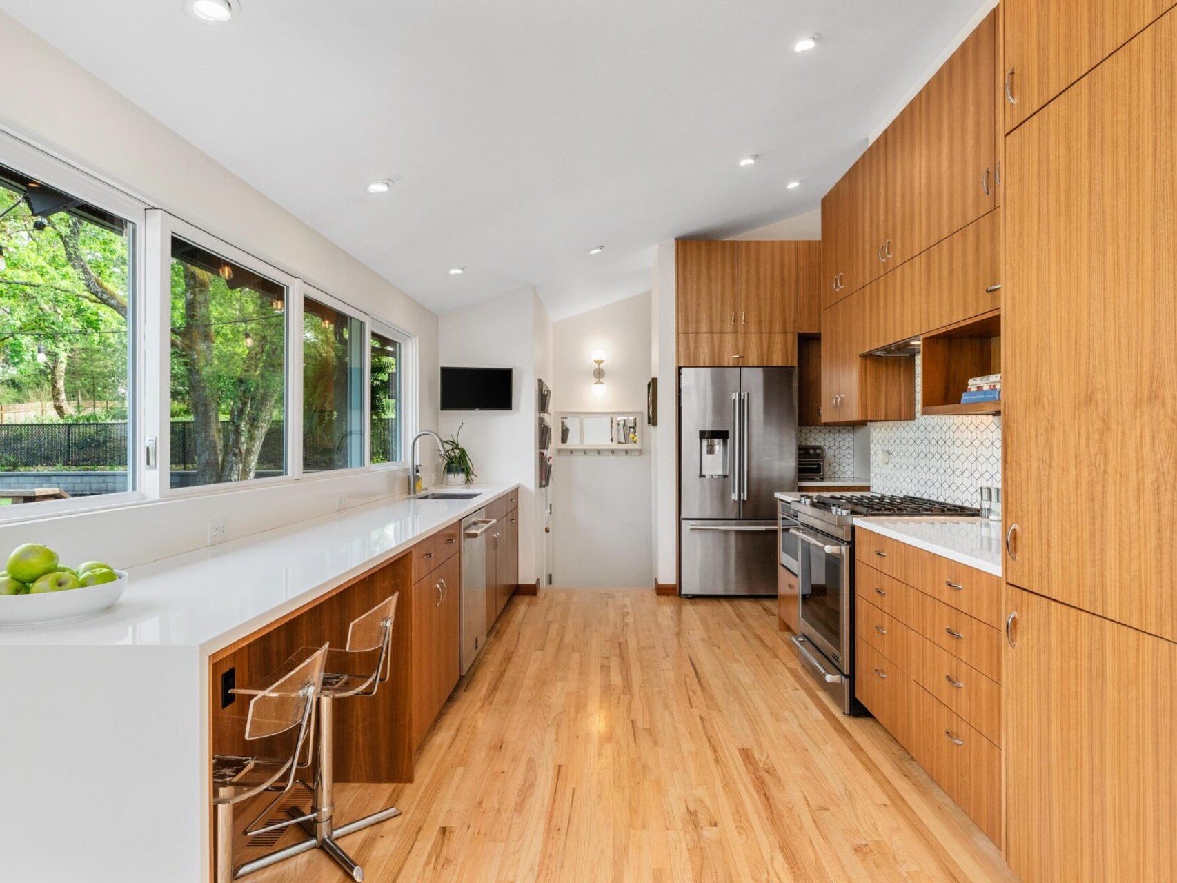 A modern kitchen with wooden cabinets and a light wood floor. It features stainless steel appliances, a white countertop with two transparent bar stools, a window overlooking greenery, and a wall-mounted TV.