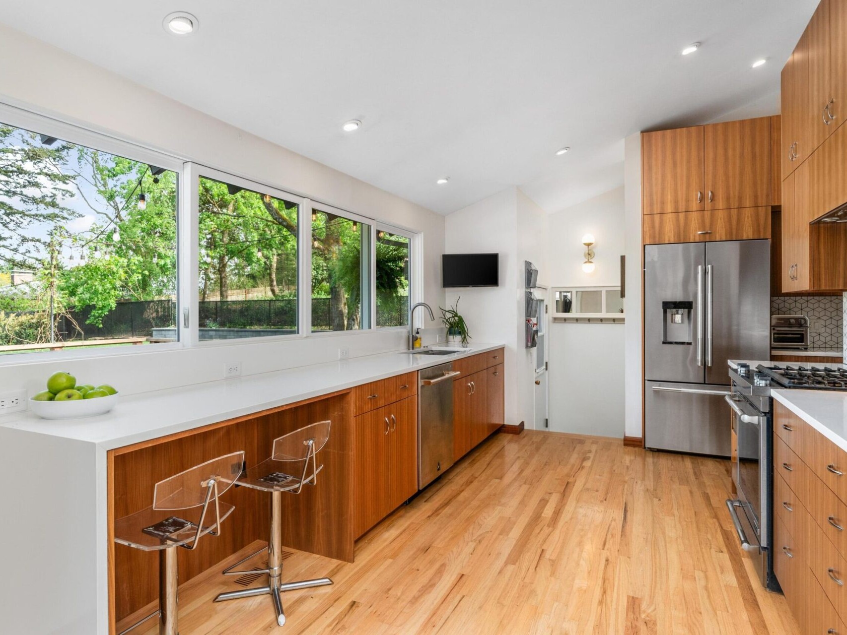 A modern kitchen with wooden cabinets, stainless steel appliances, and a white countertop. Large windows bring in natural light, and two clear barstools sit by the counter. A fruit bowl with green apples adds a touch of color.