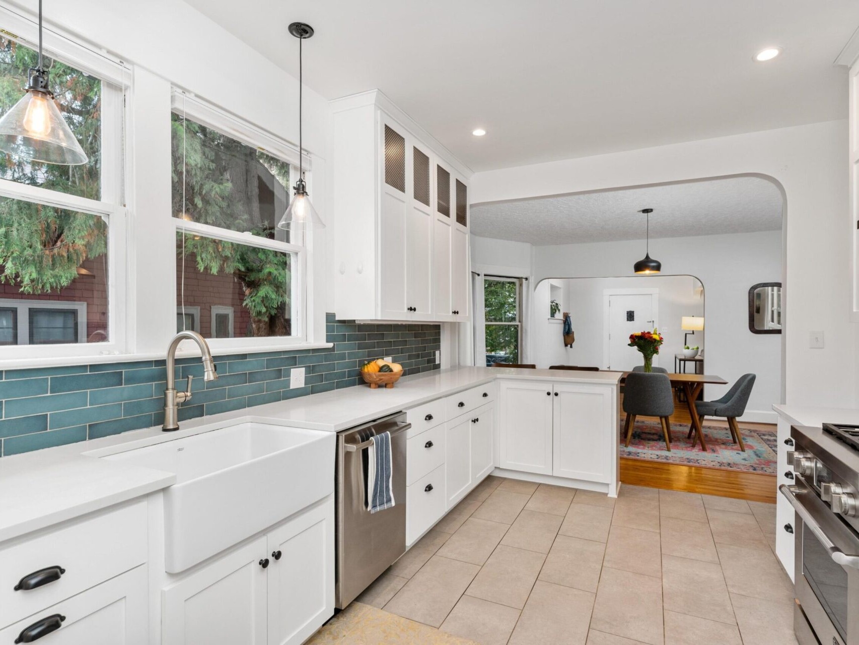 A modern kitchen with white cabinets, a farmhouse sink, stainless steel appliances, and teal subway tile backsplash. Large windows provide natural light. The adjoining dining area features a table, chairs, and a pendant light.