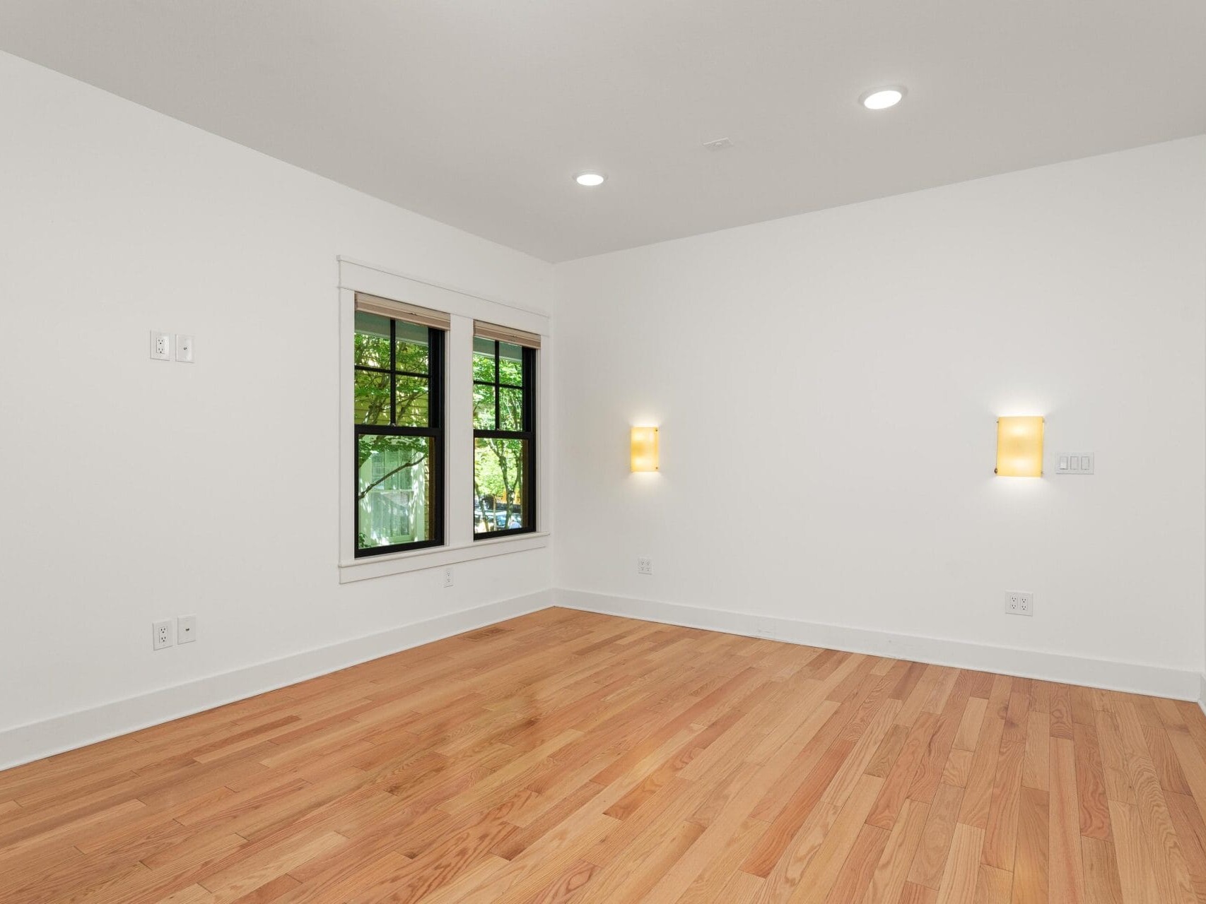 A bright, empty room with wooden floors and white walls. Two small wall lights are lit on the right, and a window on the left shows green trees outside. Recessed ceiling lights are turned on.