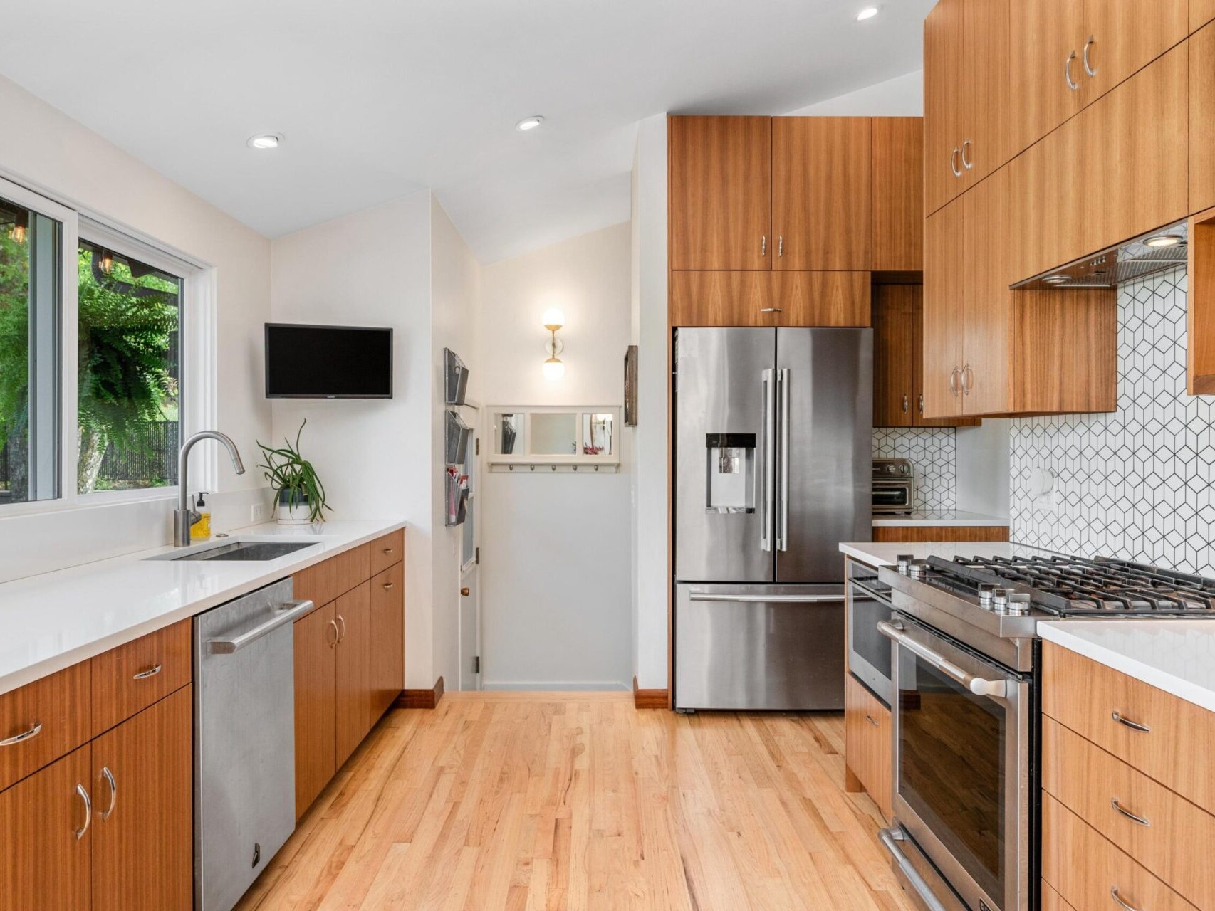 Modern kitchen with wooden cabinets, hardwood floor, stainless steel appliances, and white countertops. A window on the left lets in natural light, overlooking a green yard. A small wall-mounted screen and a plant add to the decor.