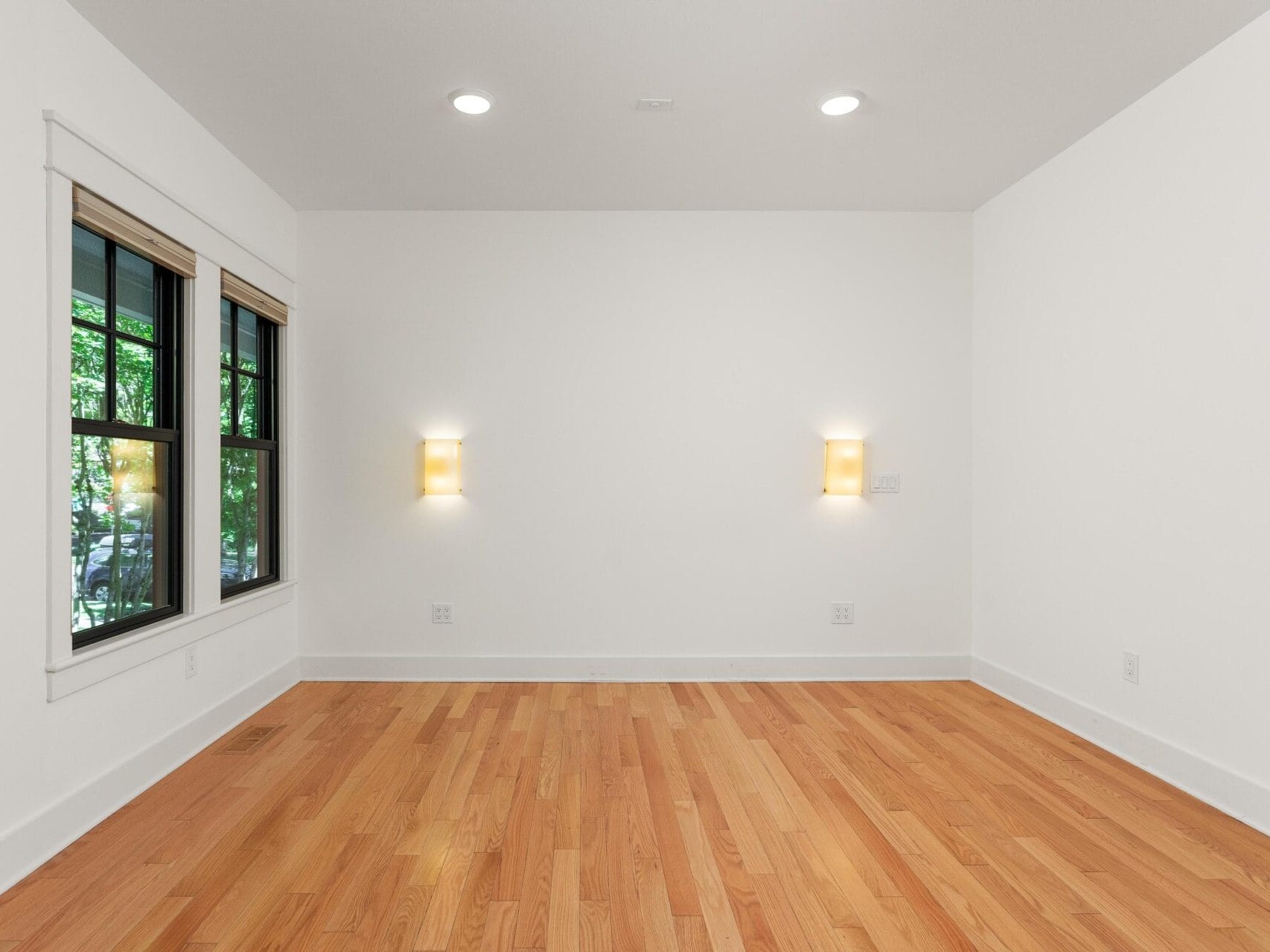 An empty room with white walls and wooden flooring. The room features two wall-mounted lights and three large windows on the left side, offering a view of greenery outside.