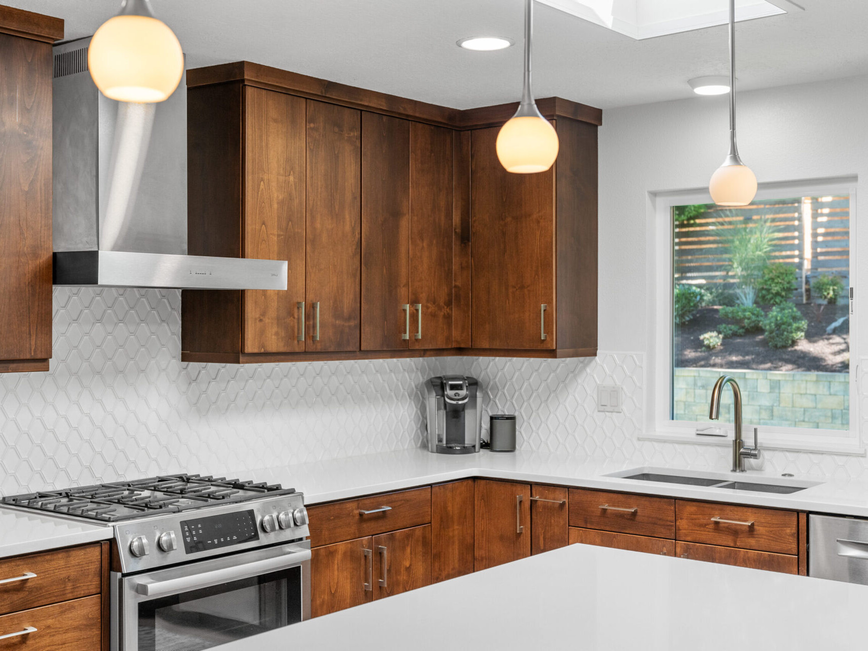 A modern kitchen with wooden cabinets, white countertops, and a stainless steel stove. Two pendant lights hang above the counter. A window near the sink provides natural light, and a coffee maker is on the counter.