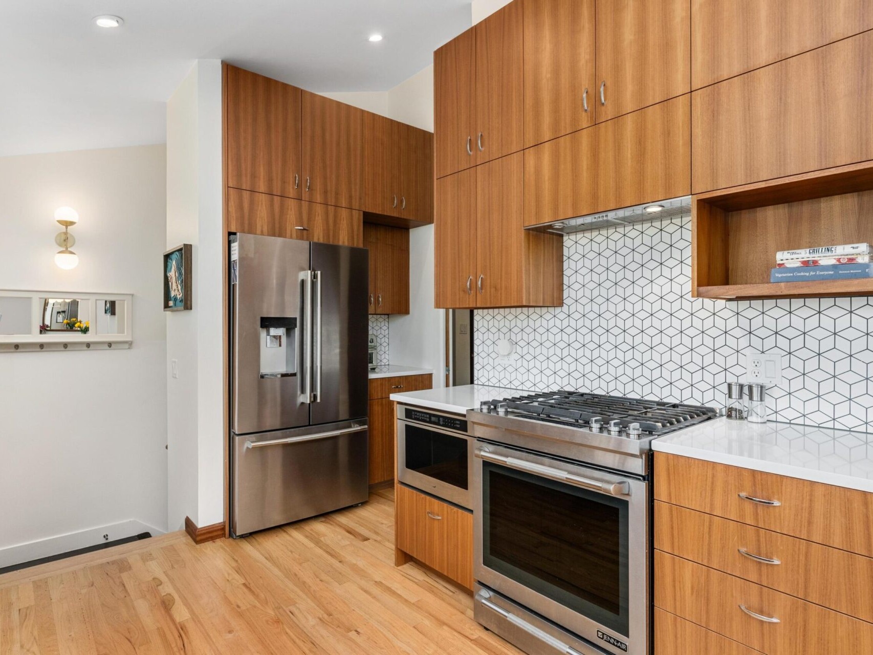 A modern kitchen with wooden cabinets, stainless steel appliances, and a hexagonal tile backsplash. There is a refrigerator, oven, and microwave, with light wood flooring and recessed lighting. Shelves display a few books and decor items.