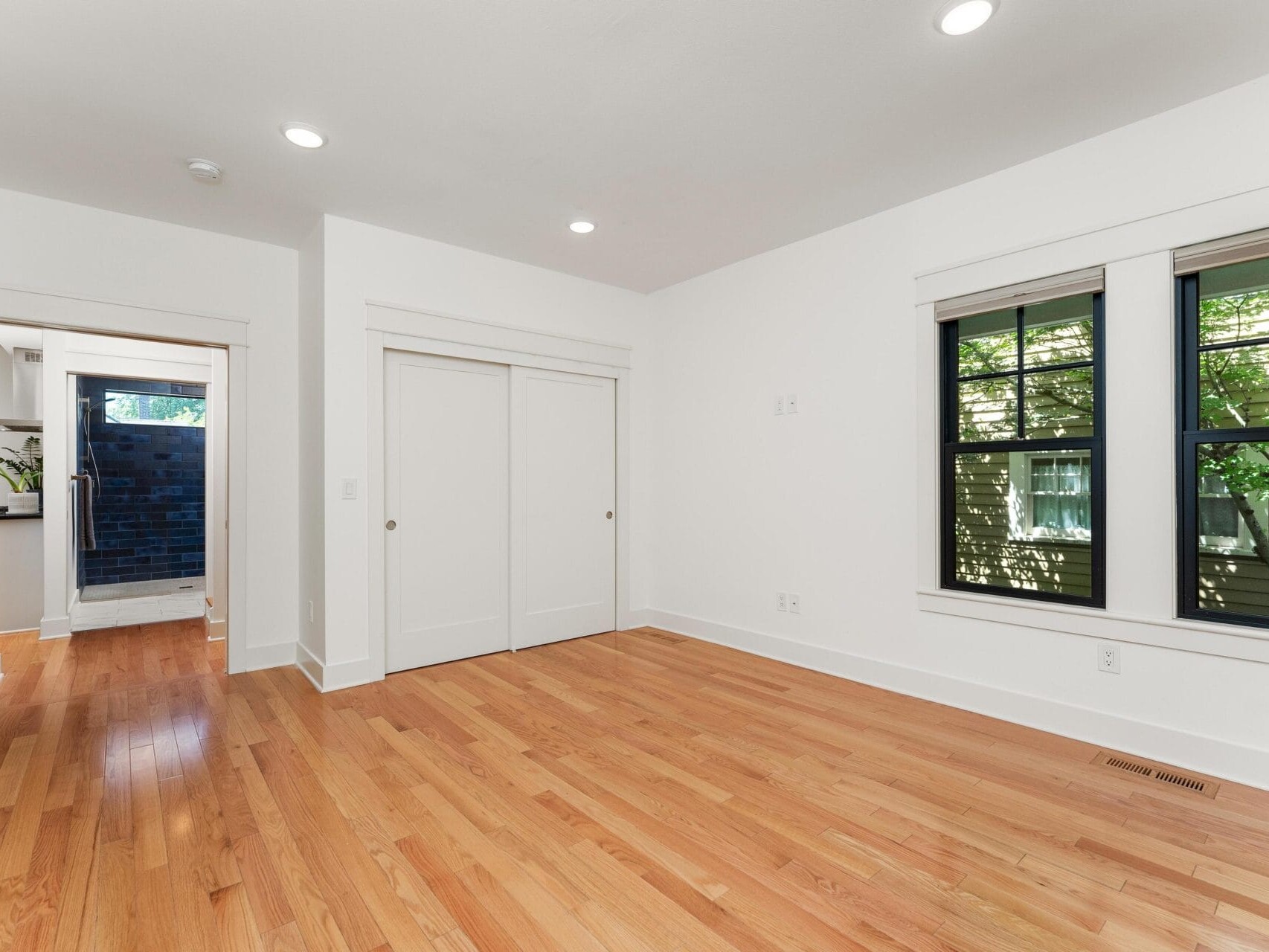 A bright, empty room with wooden floors, white walls, and recessed lighting. There are two windows on the right wall and a closet with sliding doors on the left. An open doorway leads to another room.