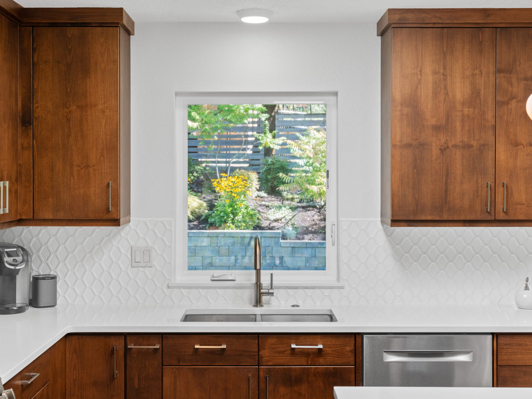 A modern kitchen features wooden cabinets, a stainless steel dishwasher, and a single-handle faucet. A window above the sink overlooks a backyard with greenery and flowers. A pendant light hangs from the ceiling, adding a warm touch.