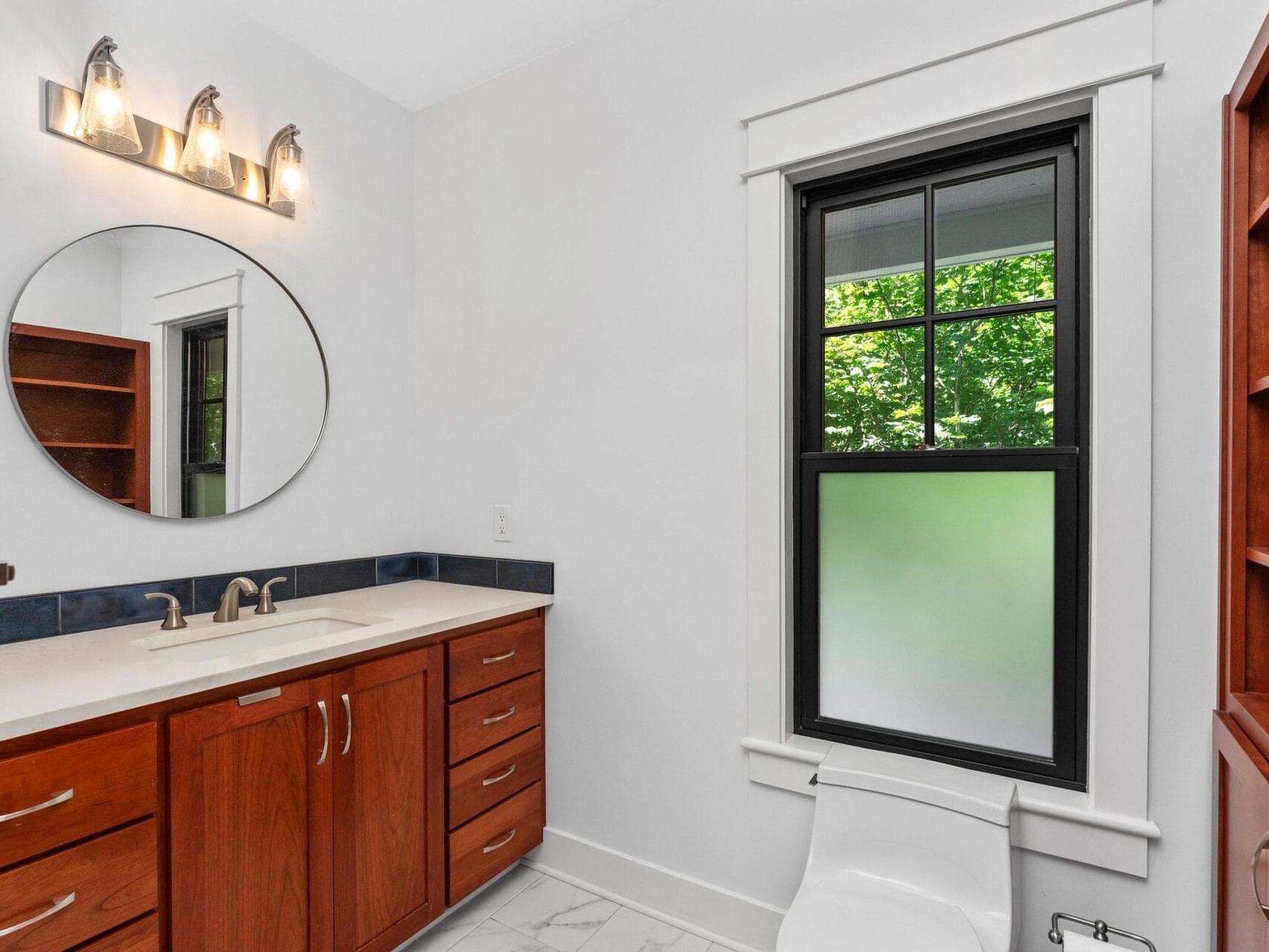 Modern bathroom with wooden cabinets, a round mirror, and a countertop with dual sinks and stainless steel faucets. A window with frosted glass provides natural light. Wall-mounted light fixtures illuminate the area.