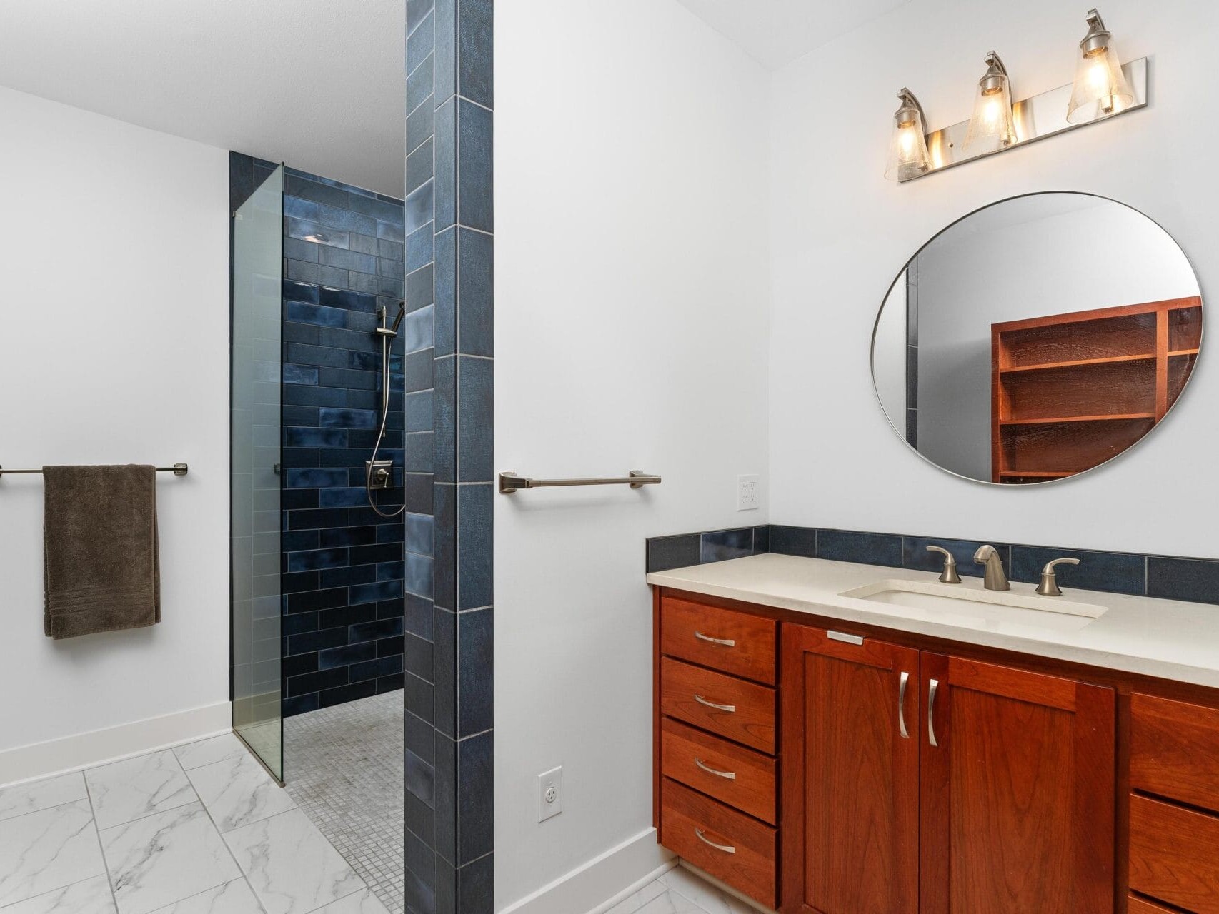 Bathroom with a wooden vanity and round mirror. The shower area has dark blue tiles and glass partition. There are white walls, a towel rack with a dark towel, marble floor, and light fixture above the mirror.