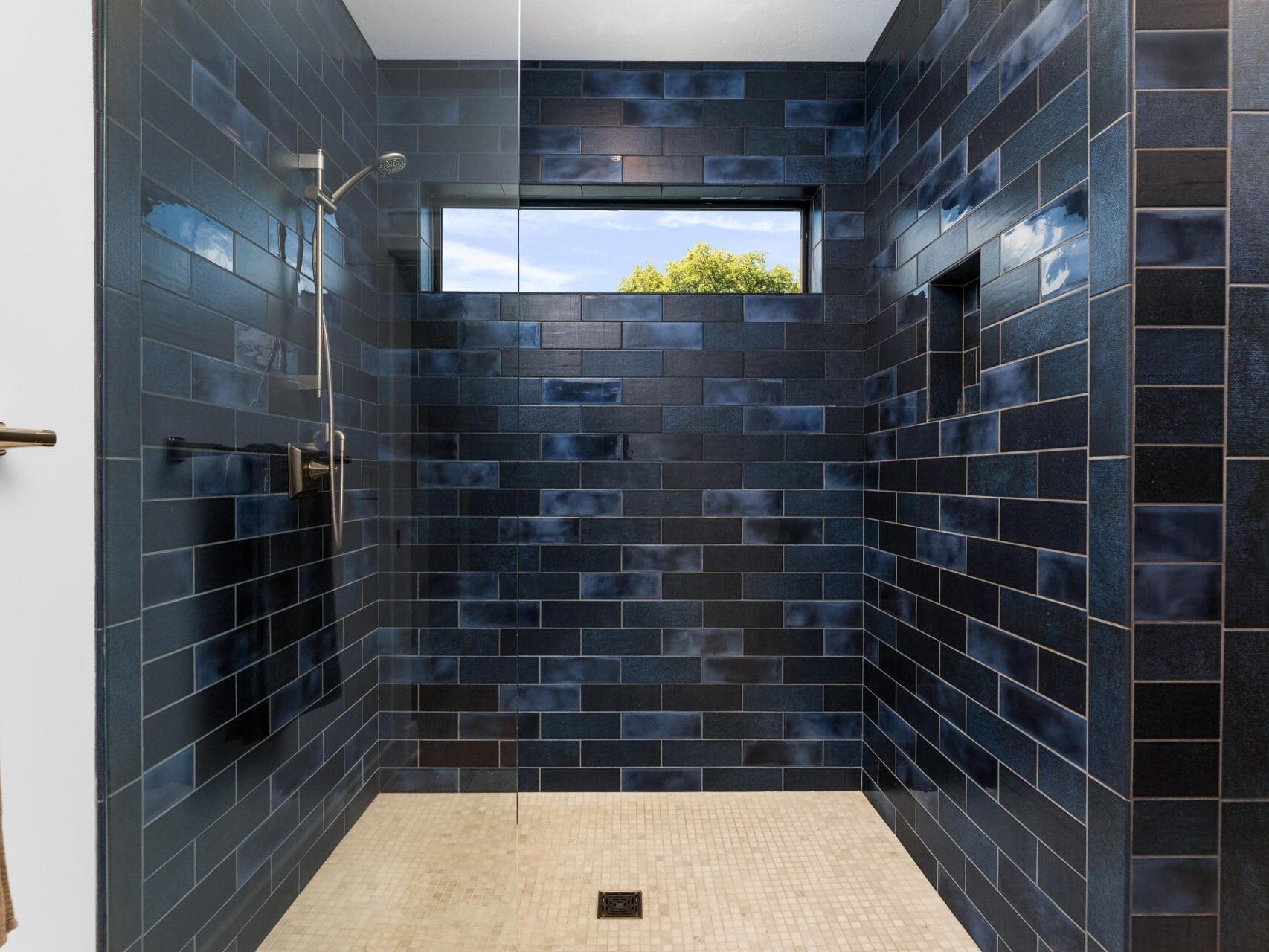A modern shower with dark blue tile walls and a glass partition. A large window brings in natural light, and theres a showerhead on the left. Beige floor tiles complement the design, and a brown towel hangs on a nearby rod.