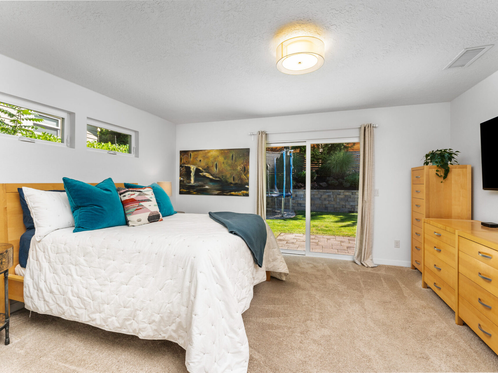 A bedroom with a large bed adorned with pillows and a quilt, wooden furniture, and a flat-screen TV. A sliding glass door leads to a brick patio with greenery outside. Neutral walls and carpeting complete the cozy space.