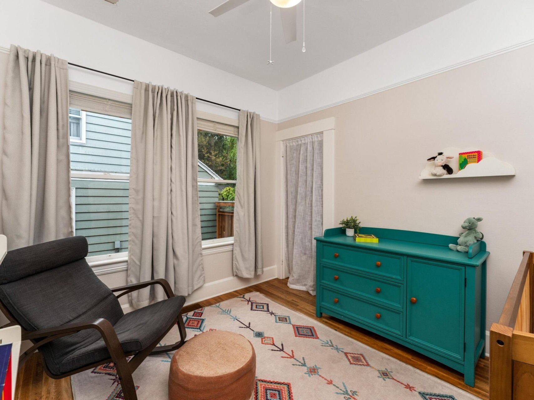A cozy nursery with a wooden crib, teal dresser, black rocking chair, and a playful rug. Soft curtains cover large windows, while a small shelf holds toys. A ceiling fan provides soft lighting.
