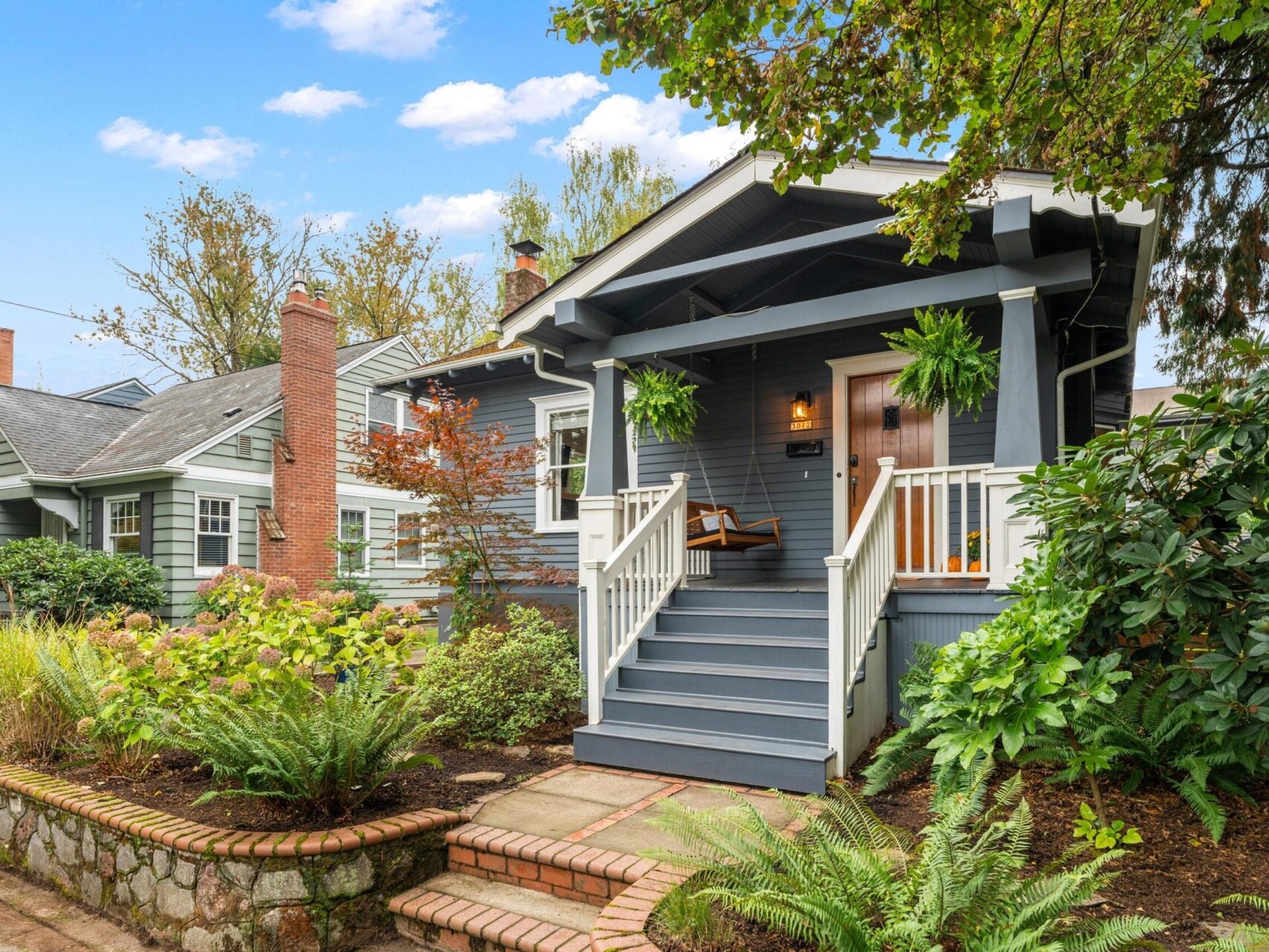 A charming blue bungalow with white trim sits under a bright sky. It features a front porch with hanging plants, wooden steps, and lush greenery in the garden. A stone pathway and brick edging enhance the landscaping.
