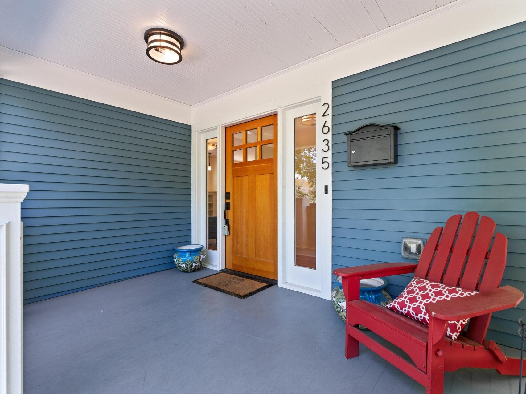 A front porch features blue siding and a wooden door with glass panels. A black mailbox is mounted on the wall. A red Adirondack chair with a patterned cushion sits beside a potted plant. The house number 2635 is displayed next to the door.