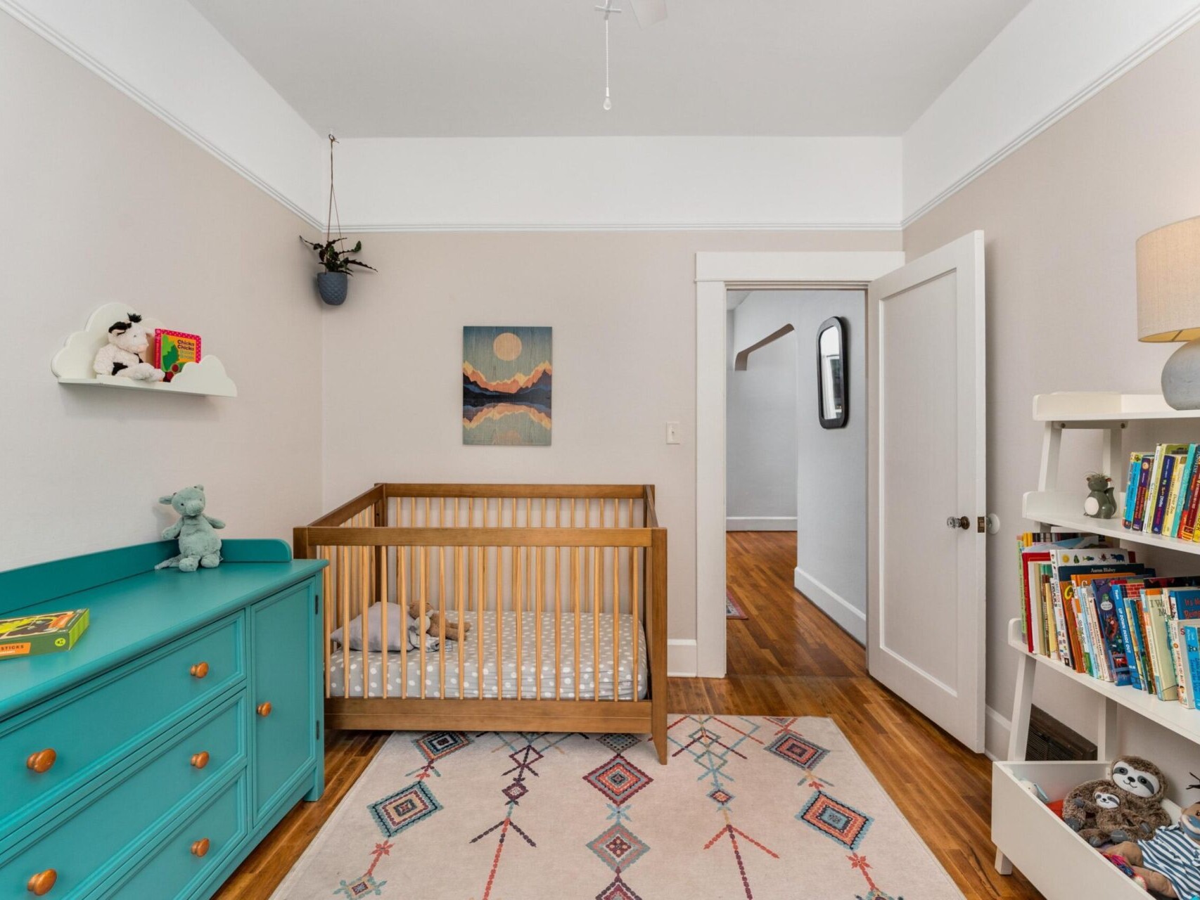 A cozy nursery features a wooden crib with a moon and mountain painting above. A teal dresser with plush toys is on the left, and a white bookshelf filled with colorful childrens books is on the right. A playful rug covers the wooden floor.