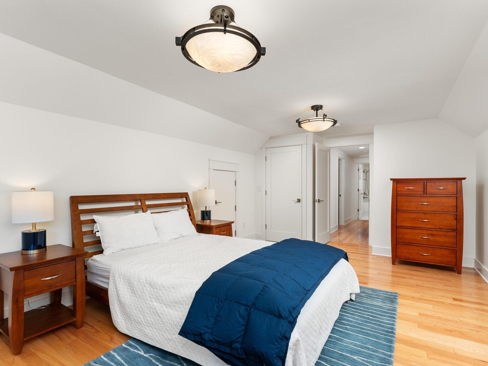 A bedroom with a wooden bed frame, blue bedding, and white sheets. Two matching wooden nightstands with lamps flank the bed. A tall wooden dresser stands to the right. The room has white walls, a ceiling light, and a hardwood floor with a blue rug.