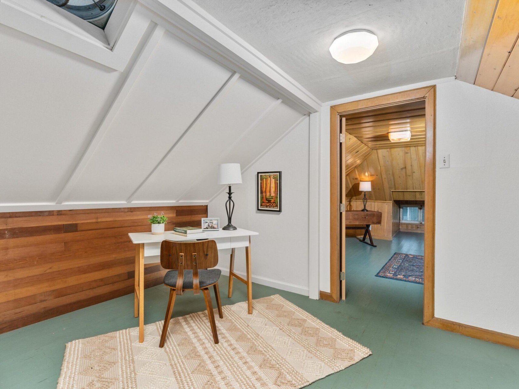 A cozy attic room with a sloped ceiling. It features a small white desk with a chair, a table lamp, and a framed picture. A doorway leads to another area, with wood paneling and a carpet visible. A rug covers part of the green floor.