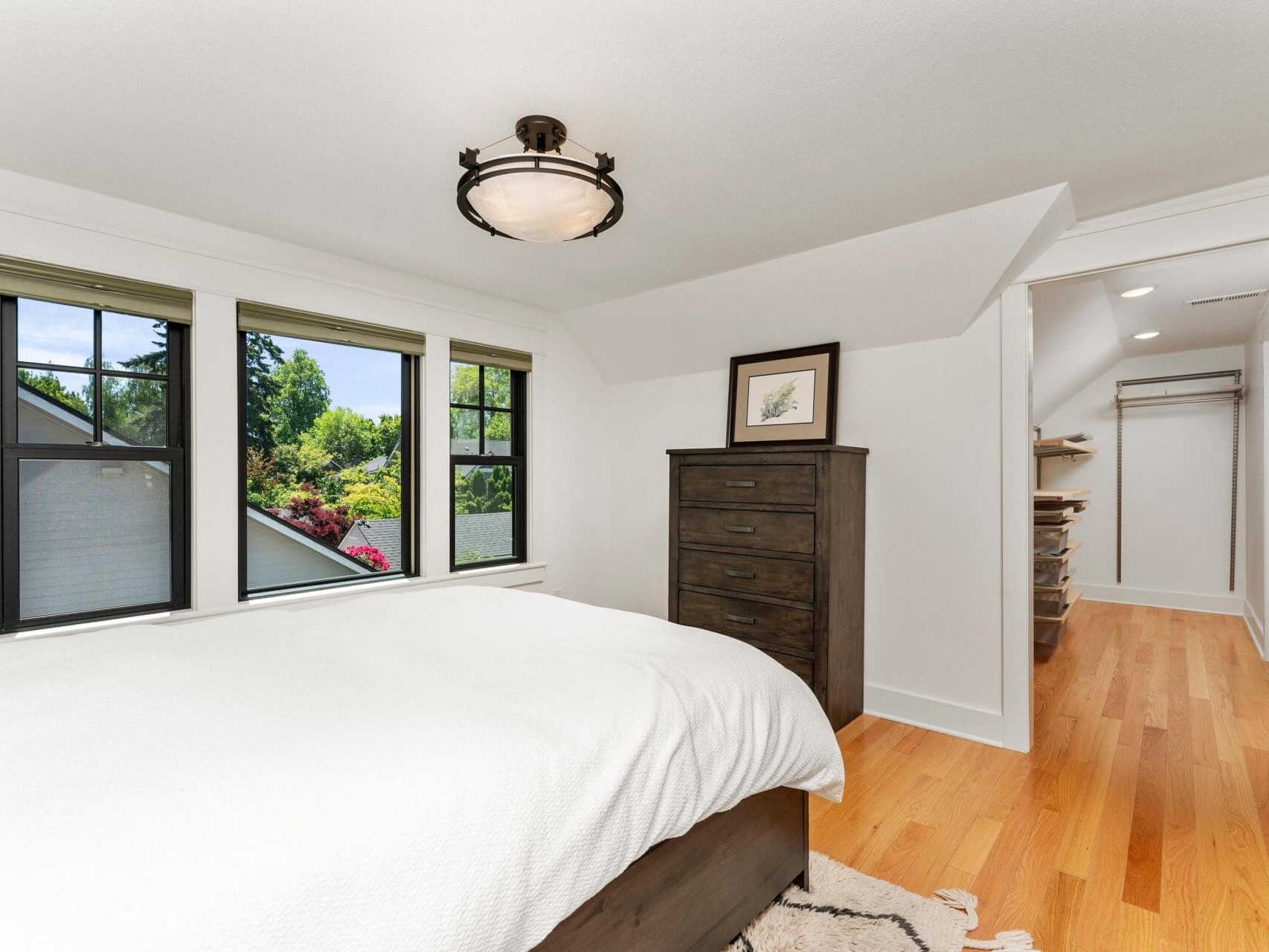 A bright bedroom with a large bed covered in a white blanket. There are three windows on the left, a dark wooden dresser, and an open closet with wooden floors visible through a doorway. Natural light fills the room.