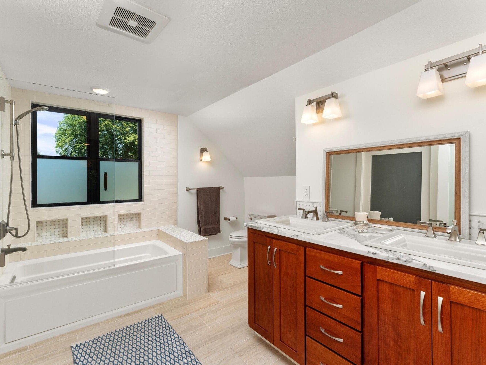 A modern bathroom with a bathtub and shower combo near a large frosted window. A double-sink vanity with a wooden cabinet and a large mirror is on the right. The room has beige tile flooring and contemporary light fixtures.
